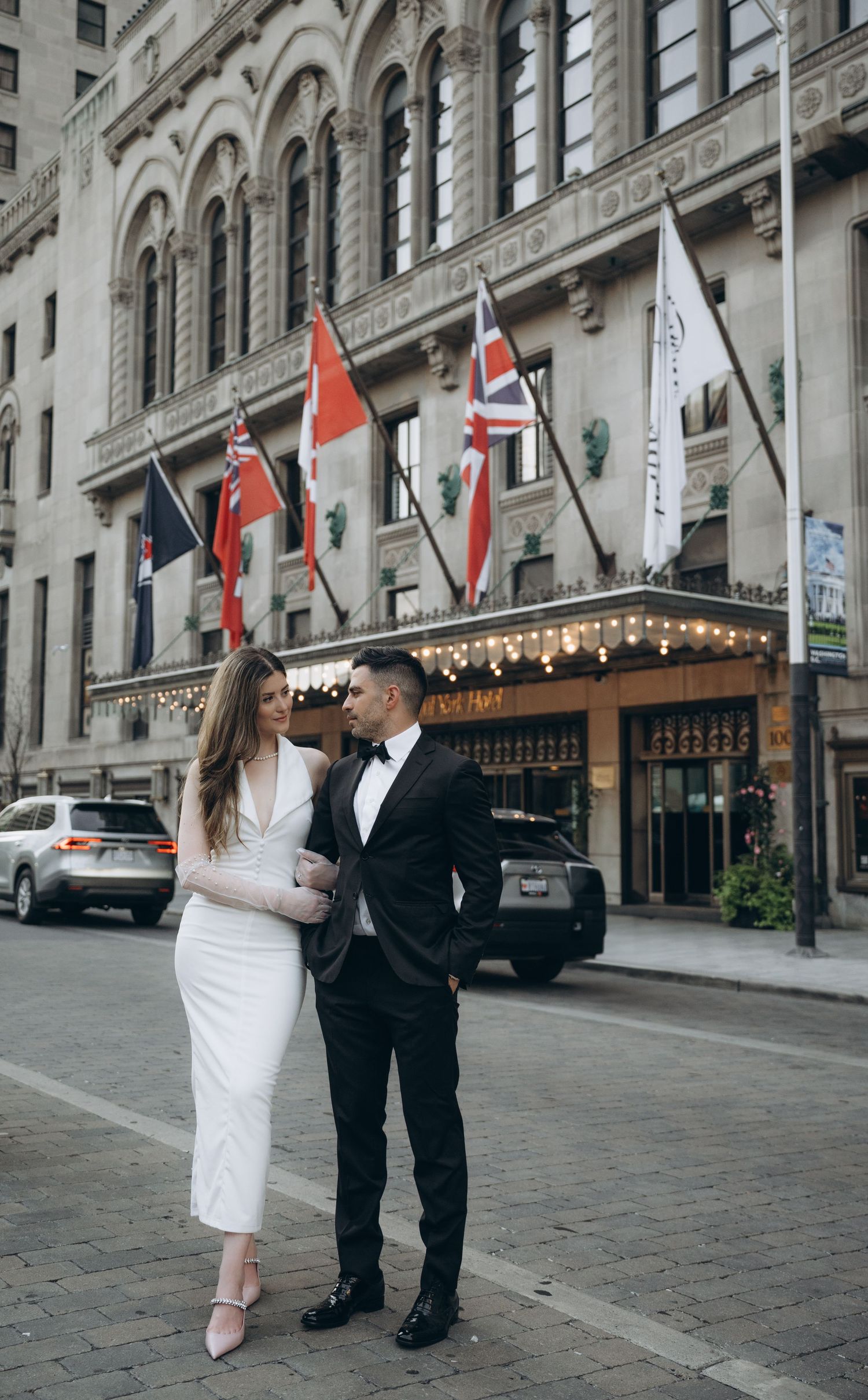 A couple in wedding attire shares a romantic moment on a historic cobblestone street with ornate building facades and flags.