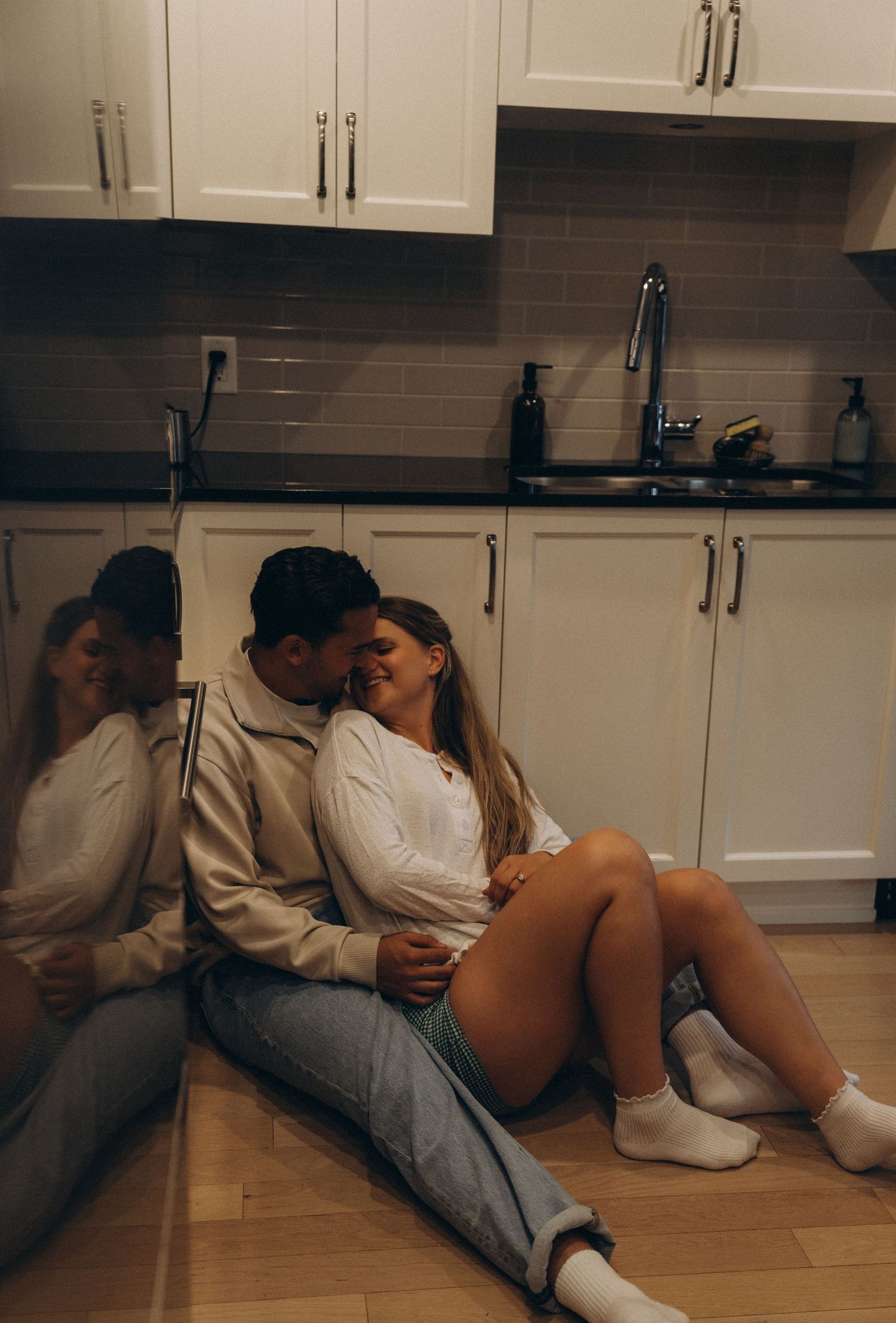 Couple sitting together intimately on kitchen floor with modern white cabinets and dark tile backsplash in background.