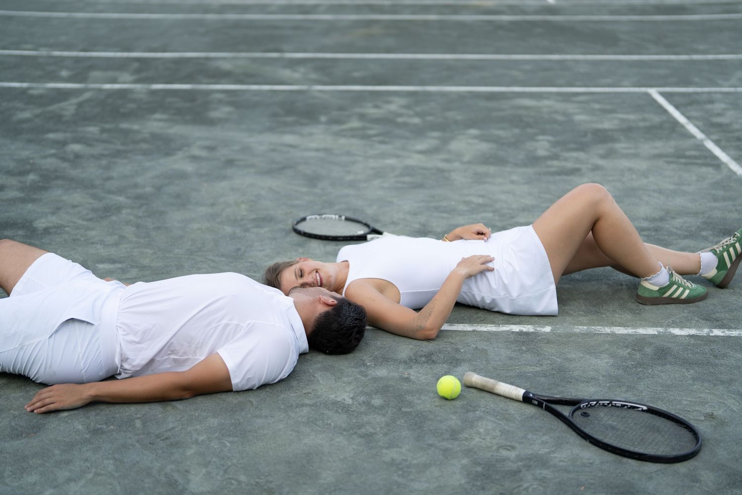 Two people lay side by side on a tennis court with rackets and tennis balls nearby.