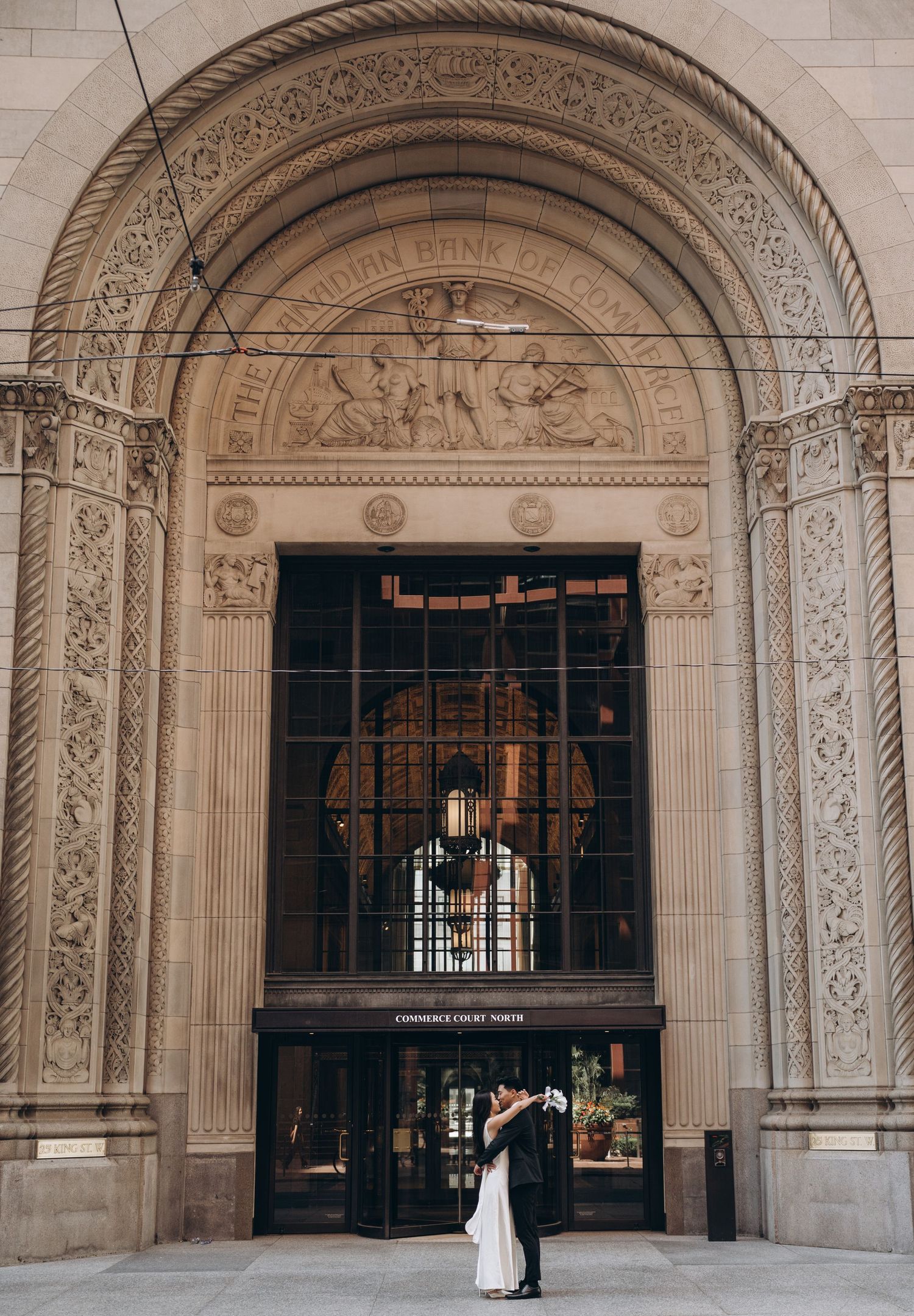 Elegant wedding couple embraces beneath ornate stone archway entrance of historic building with decorative reliefs.
