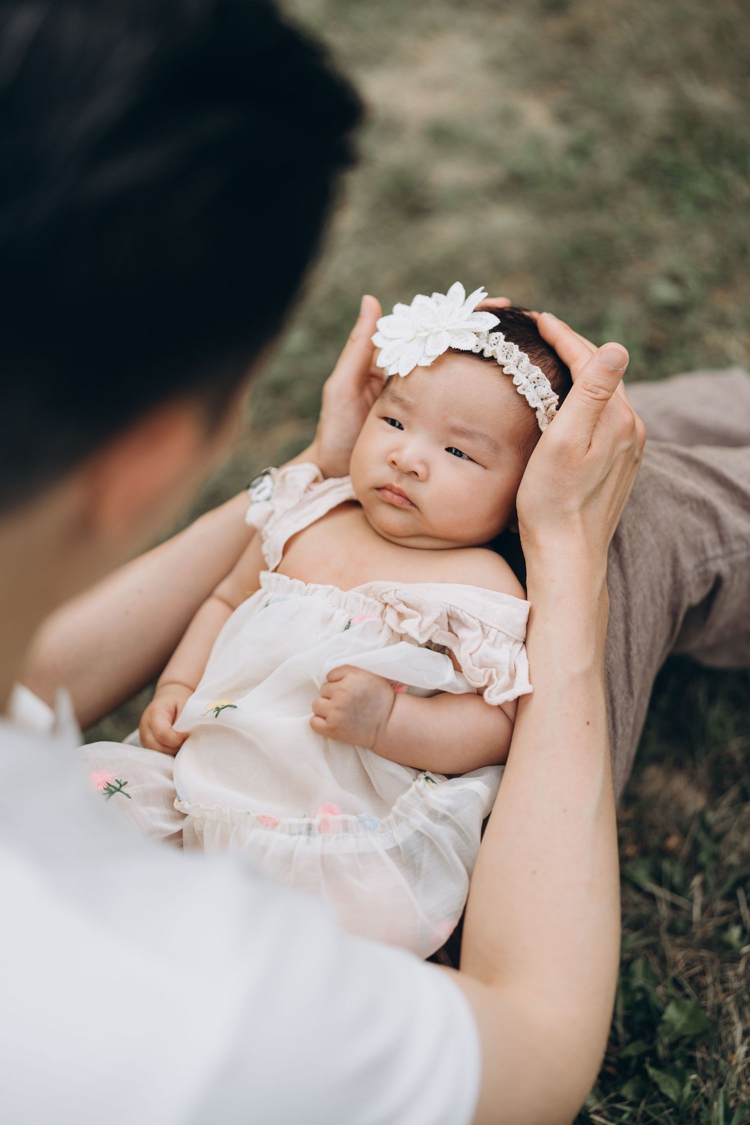 A baby wearing a white dress and floral headband lies in someone's arms on grass outdoors.