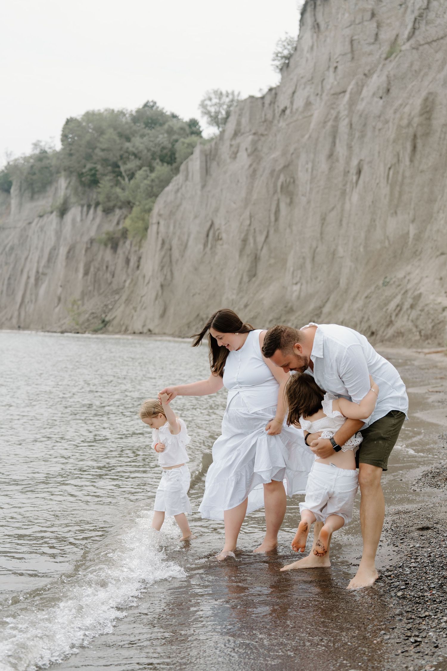 Family in white outfits splashing and playing together at the water's edge near scenic cliffs.