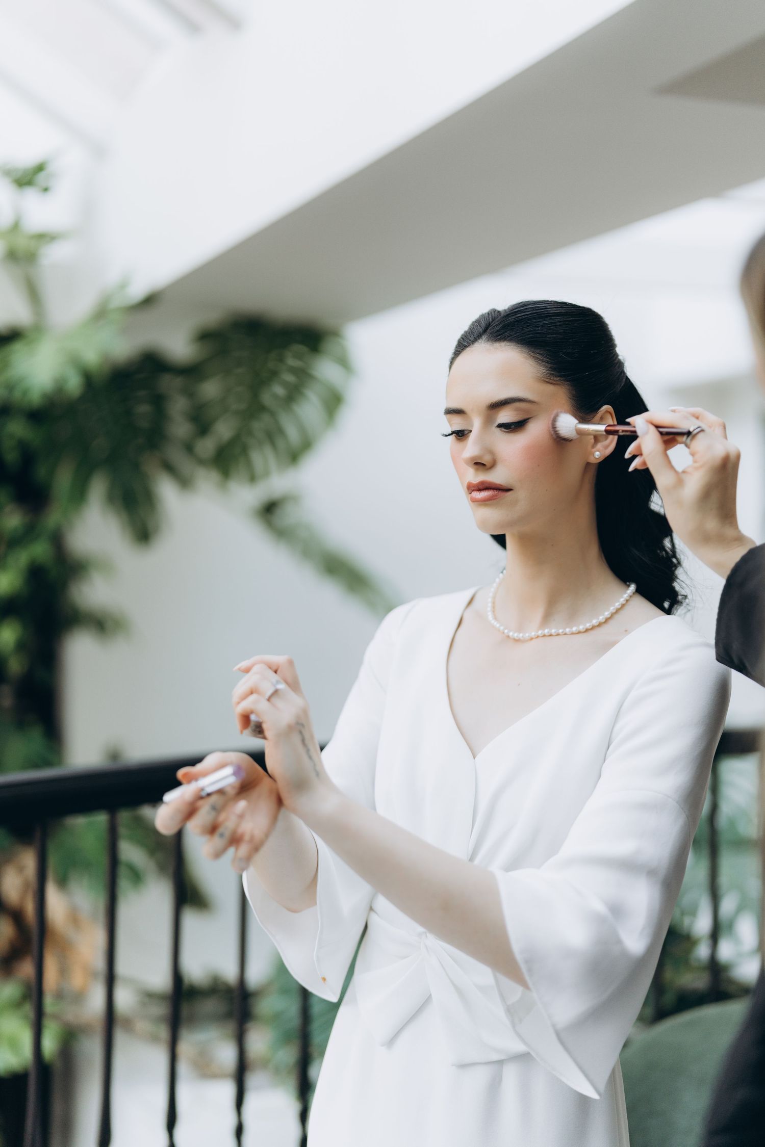 A woman in a white dress prepares for a special occasion while getting her makeup done in a bright indoor setting.