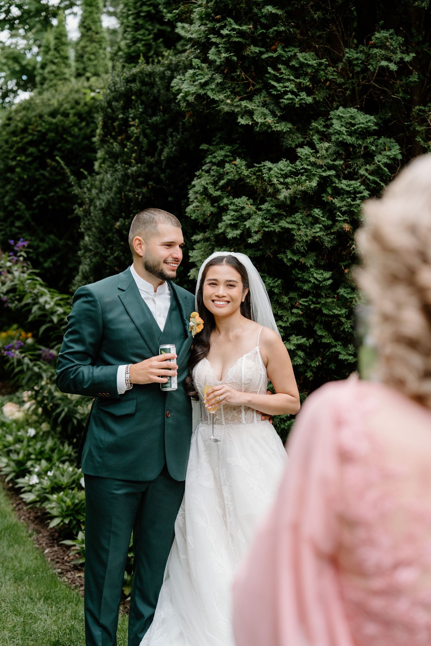 A couple celebrates with drinks during their outdoor wedding reception surrounded by lush greenery.