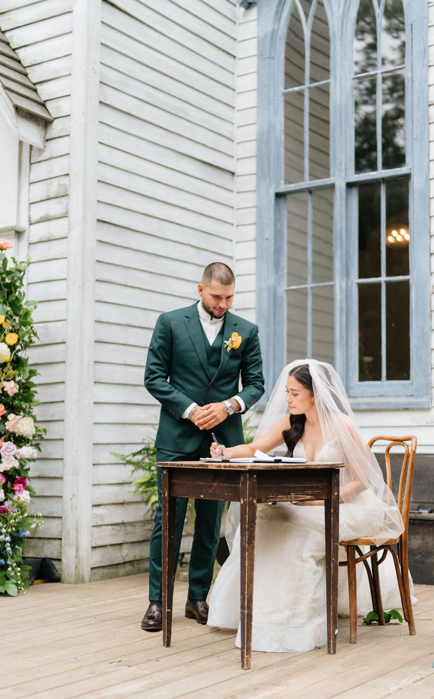 A couple signs their marriage license at a wooden table on a white church porch decorated with floral arrangements.