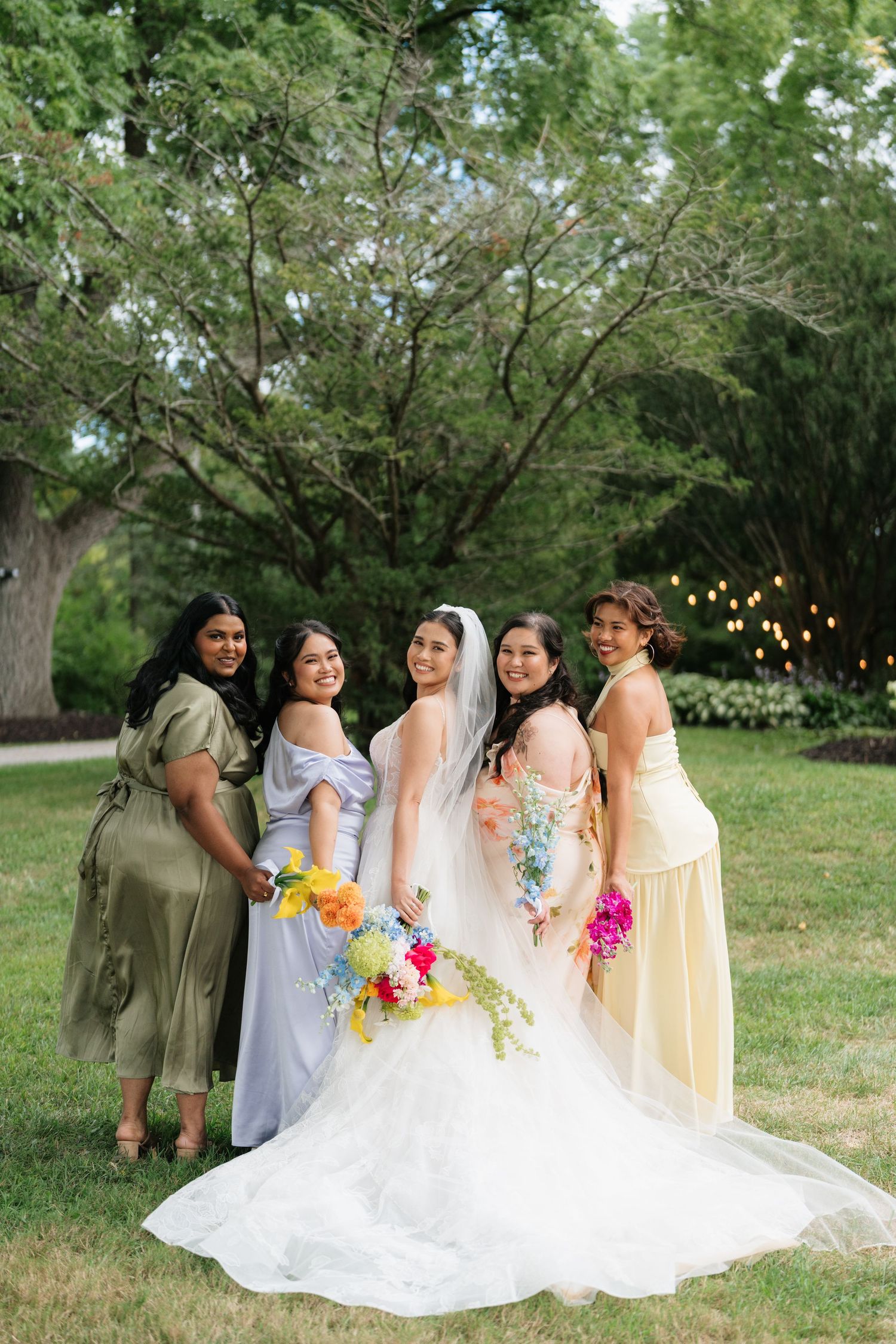 A bridal party in pastel dresses and a white wedding gown poses together outdoors under green trees with string lights.