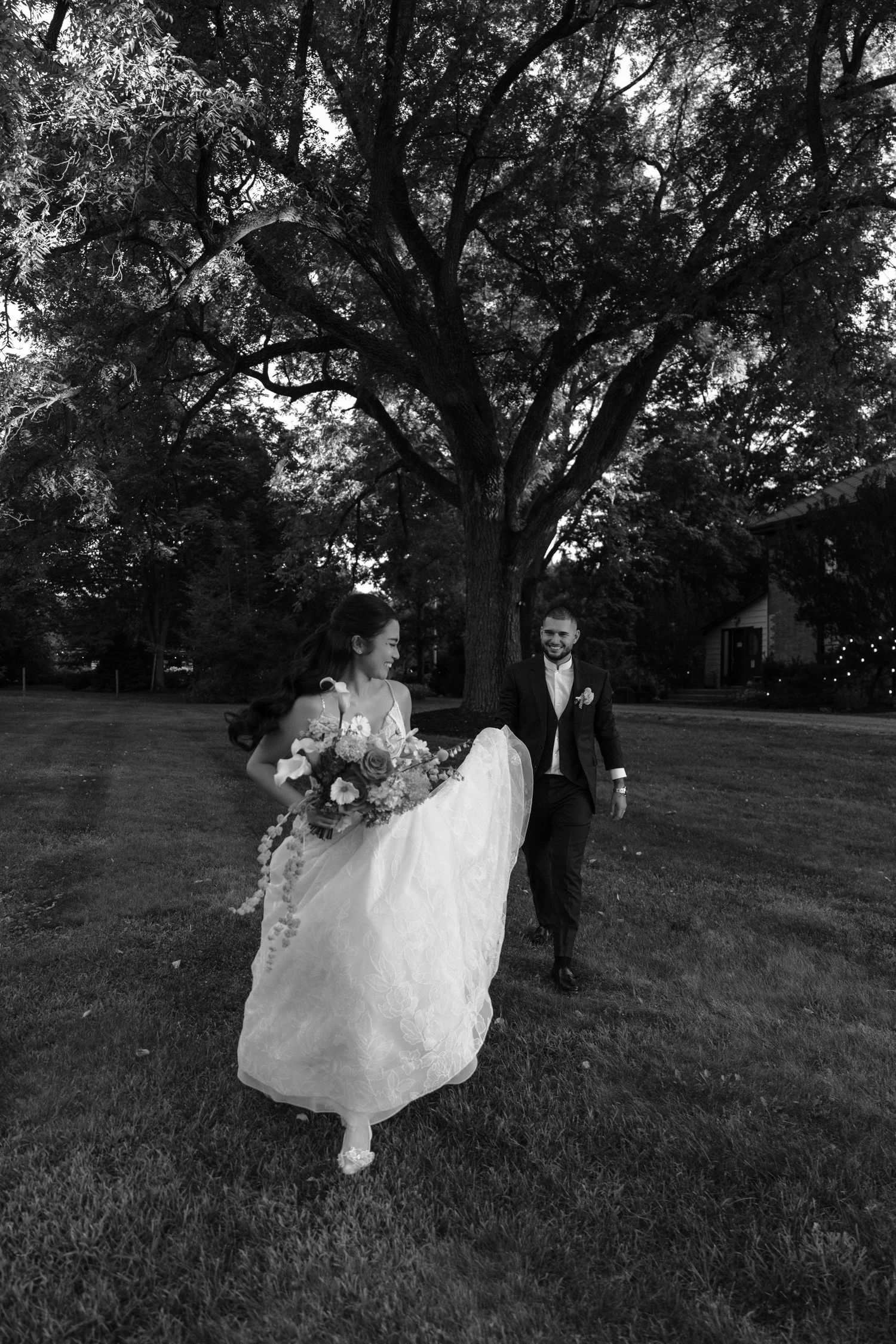 Black and white wedding photo of couple walking through grassy field with trees.