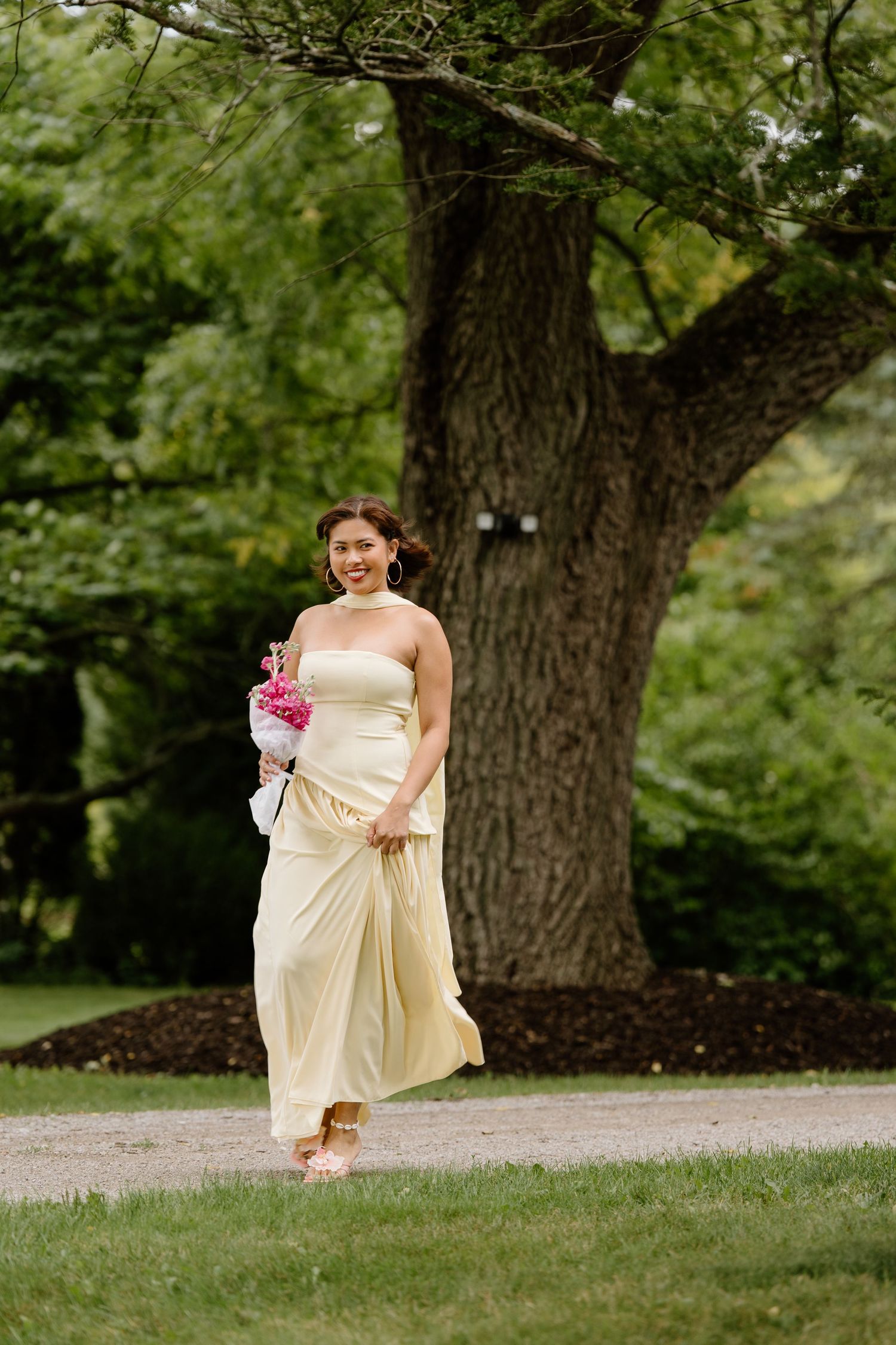 A person in a cream-colored gown holds a pink bouquet while walking on grass near a large tree in a garden setting.