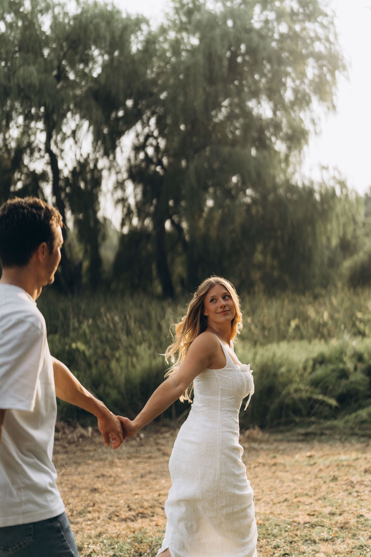 A couple in white outfits holds hands while walking through a sunlit field with trees in the background.in Toronto by AZ Photo Studio.