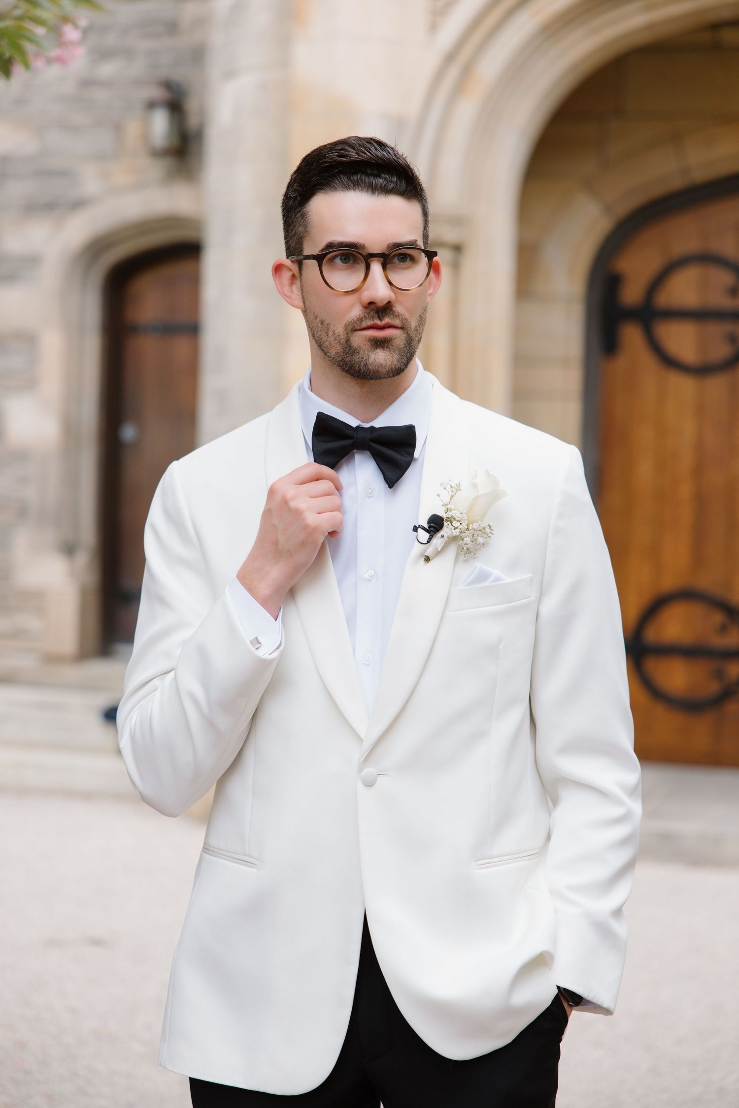 A well-dressed person in a white tuxedo jacket, black bow tie and glasses stands in front of an elegant stone archway.