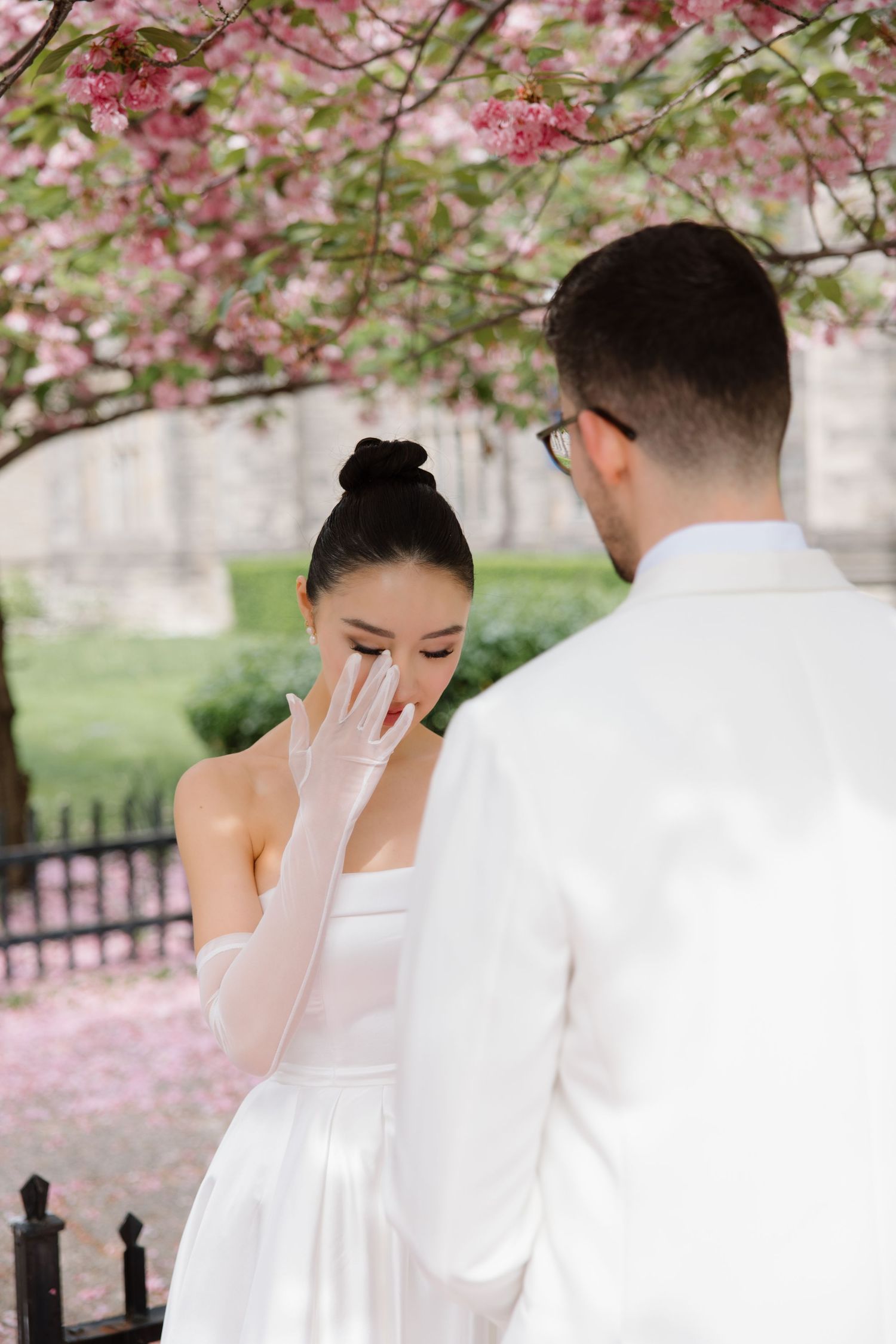Two people in wedding attire share an emotional moment under blooming cherry blossoms on a spring day.