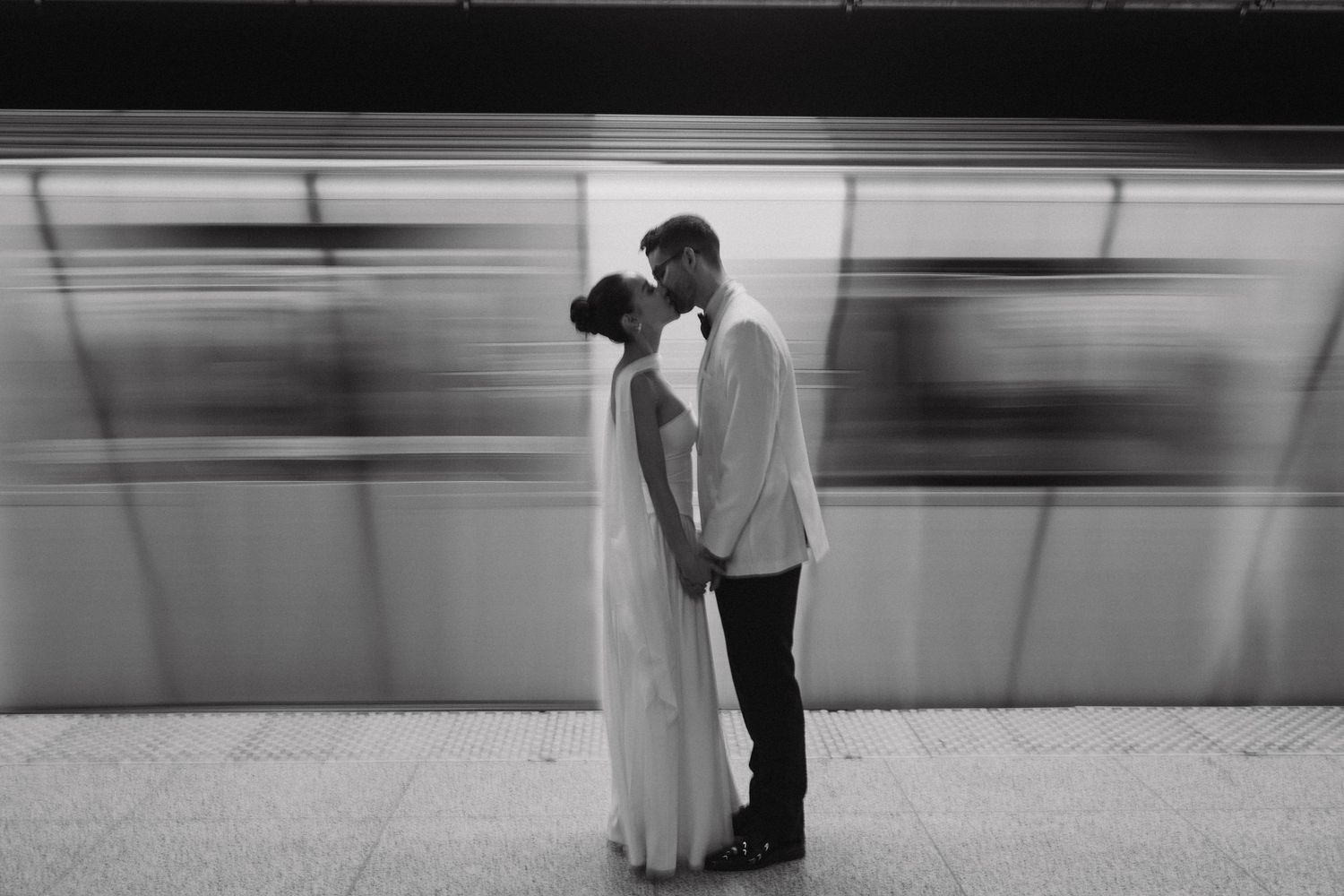 A romantic couple shares a kiss on a subway platform with a blurred moving train in the background.