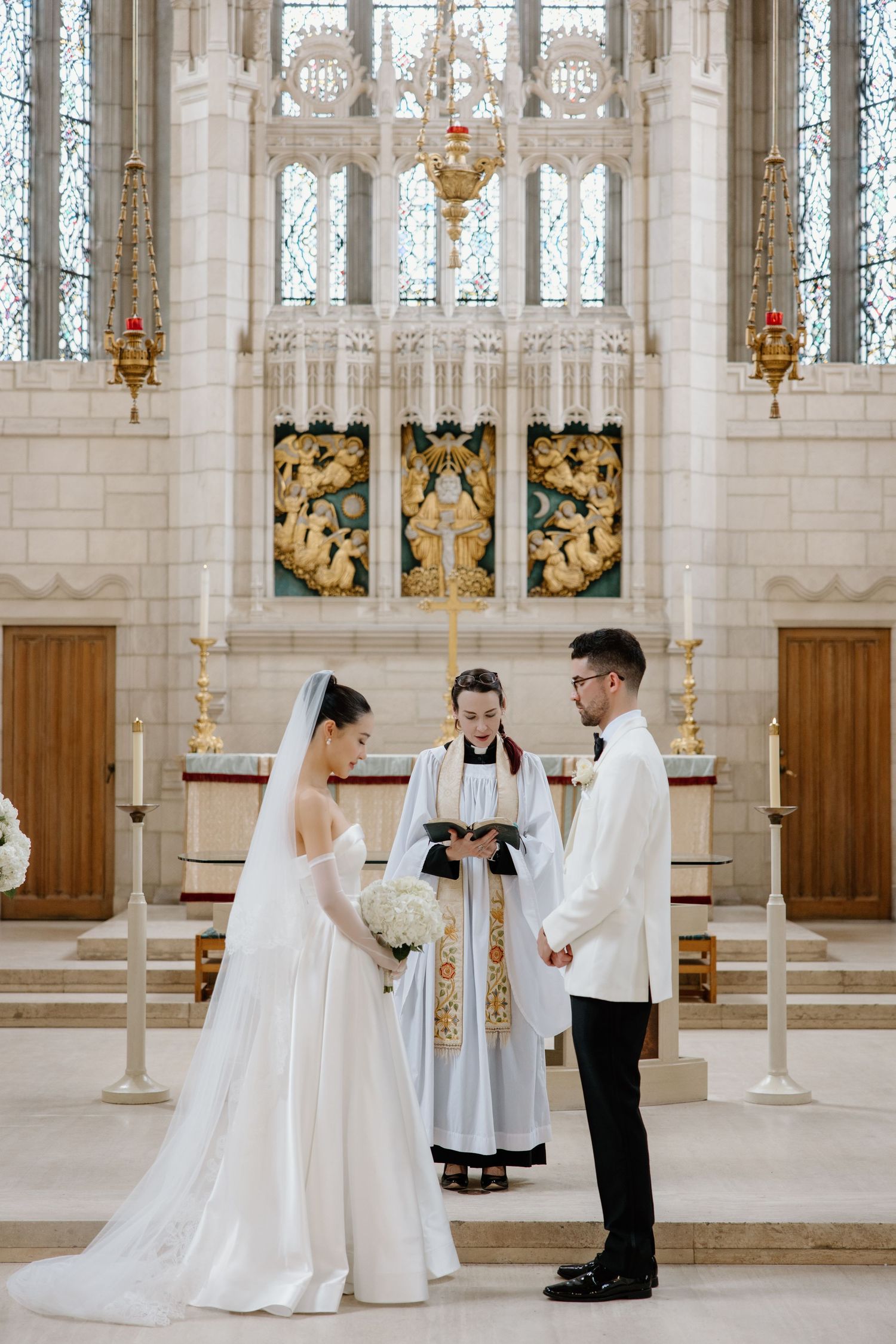 A wedding ceremony takes place in a grand Gothic church with ornate stained glass windows and golden decorative panels.