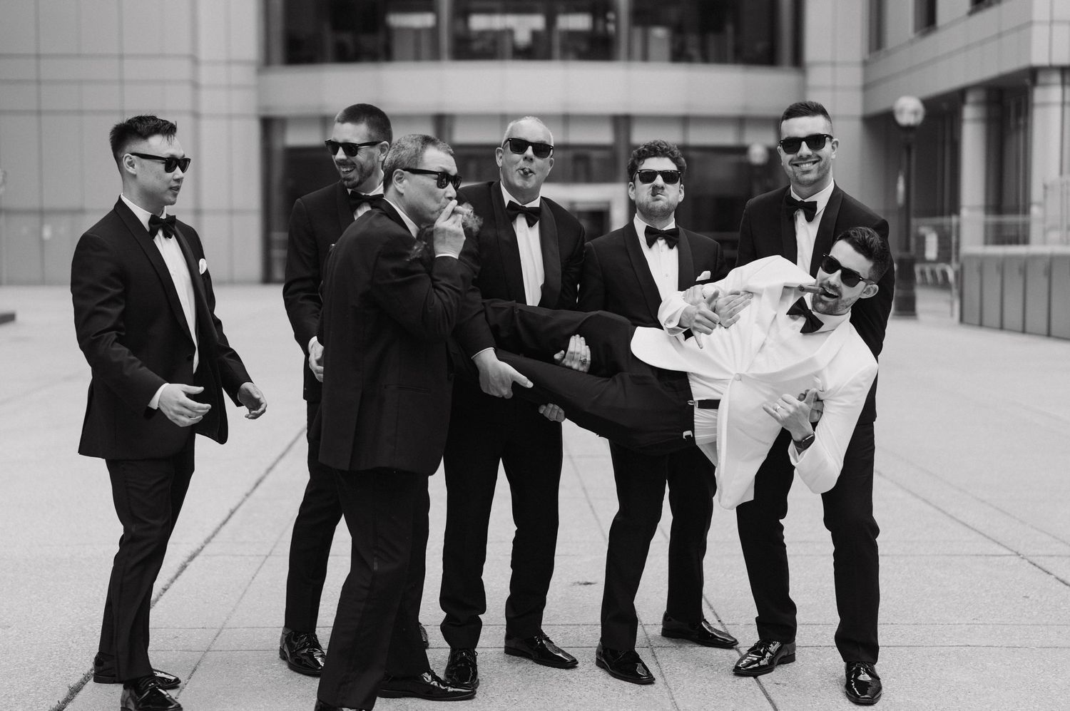 Groomsmen in black suits carry another person wearing white in a playful pose outside a modern building.