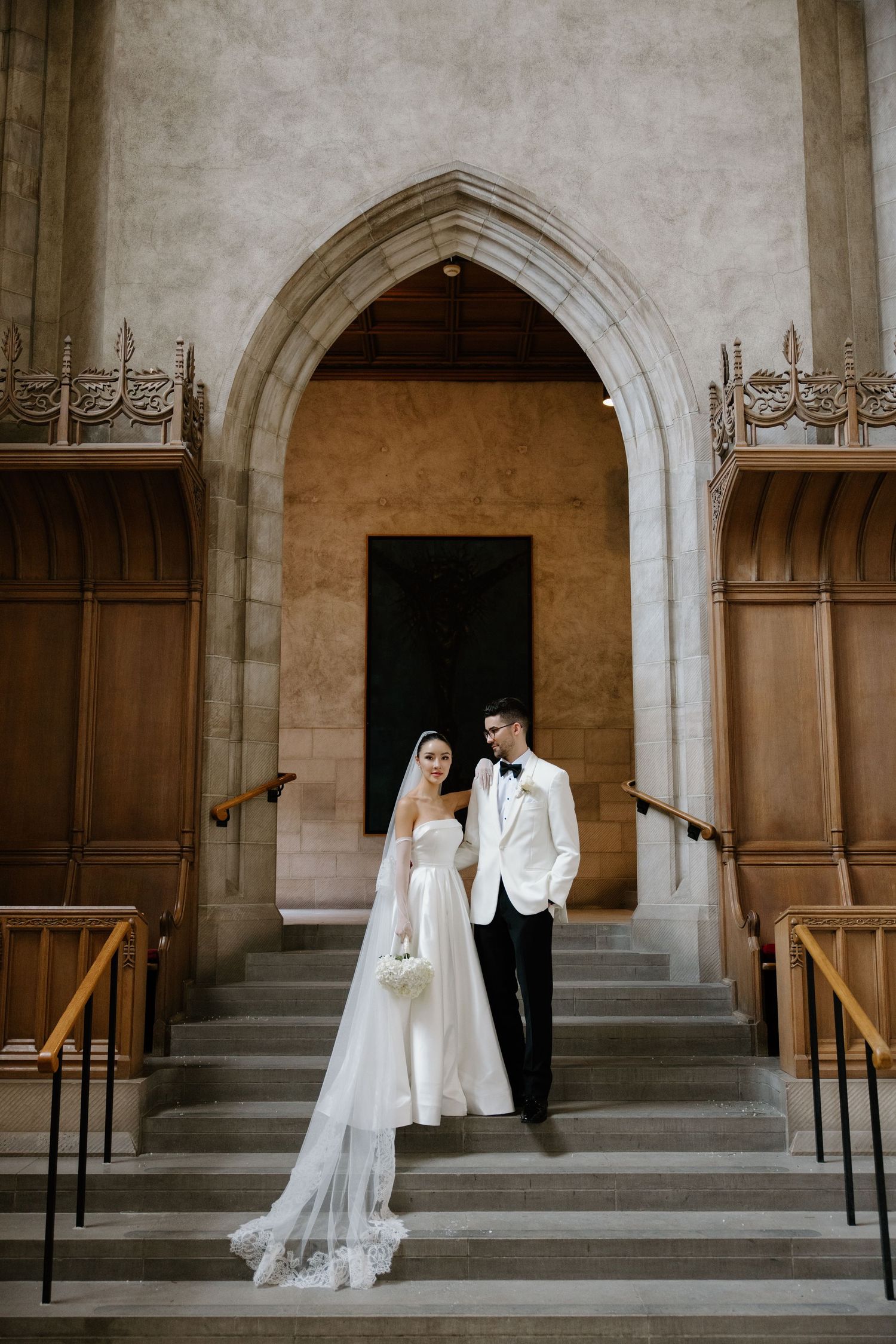 A bride in a flowing white dress and groom in white tuxedo pose elegantly on church steps beneath a gothic archway.