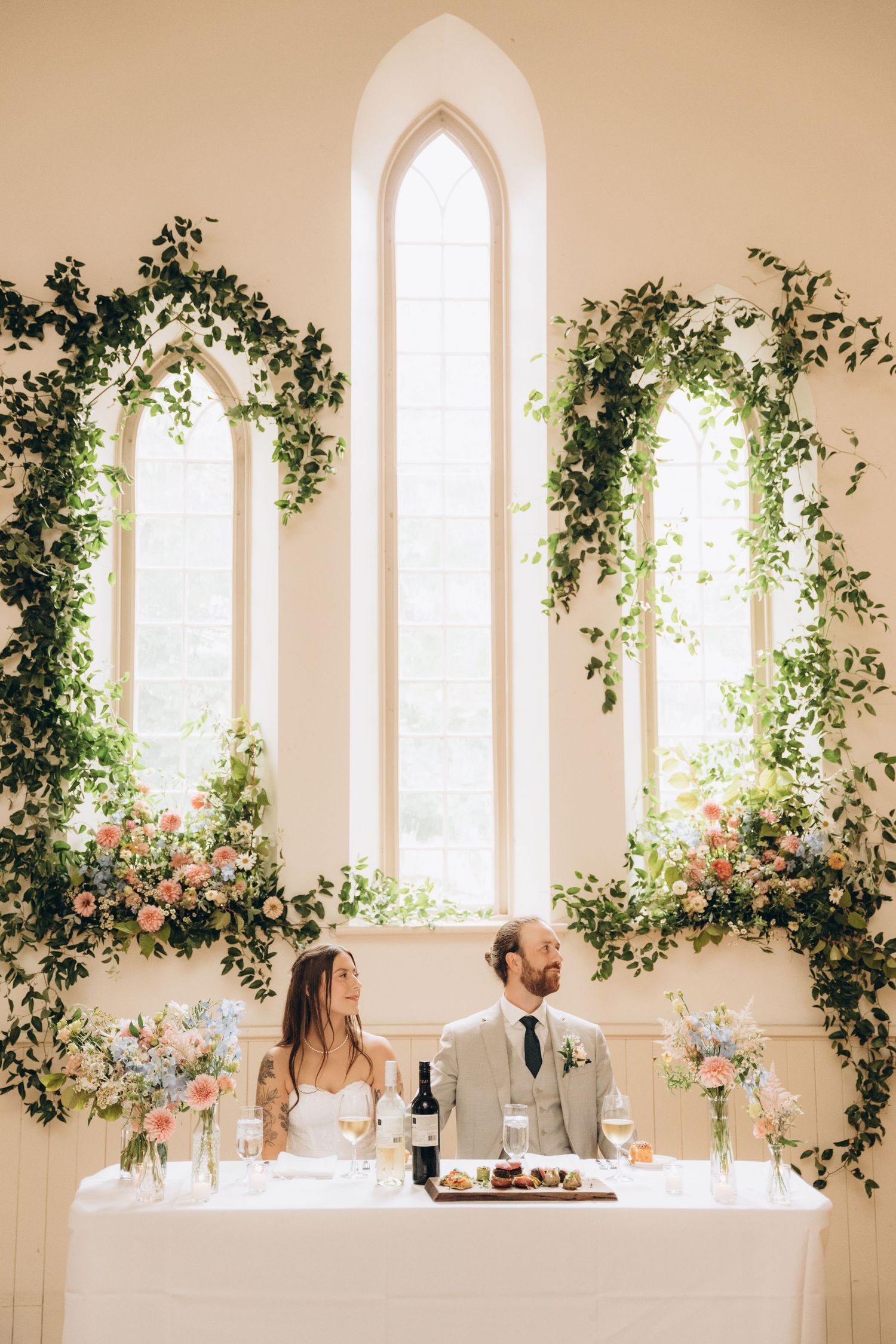 Wedding reception decor features elegant white arched windows adorned with lush green vines and pink floral arrangements.