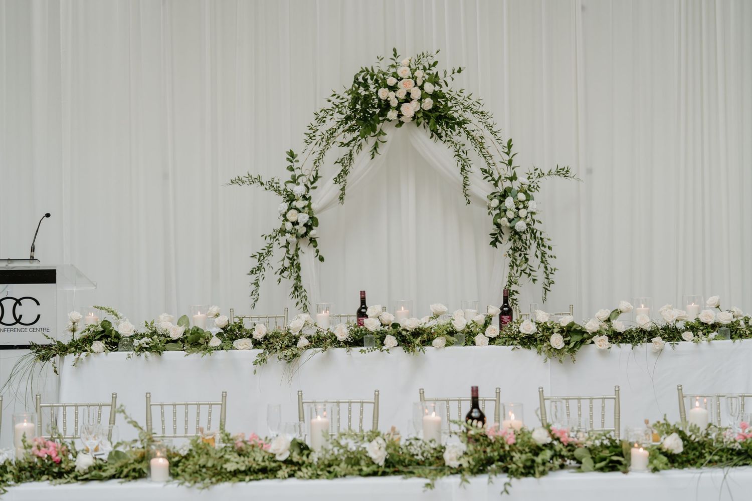 Elegant white floral arch and table decorations for a wedding reception with white chairs and greenery garlands.