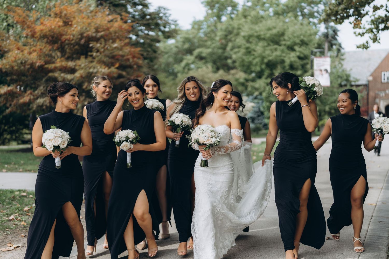 Wedding party in black dresses and formal attire walking together outdoors on a sunny day.