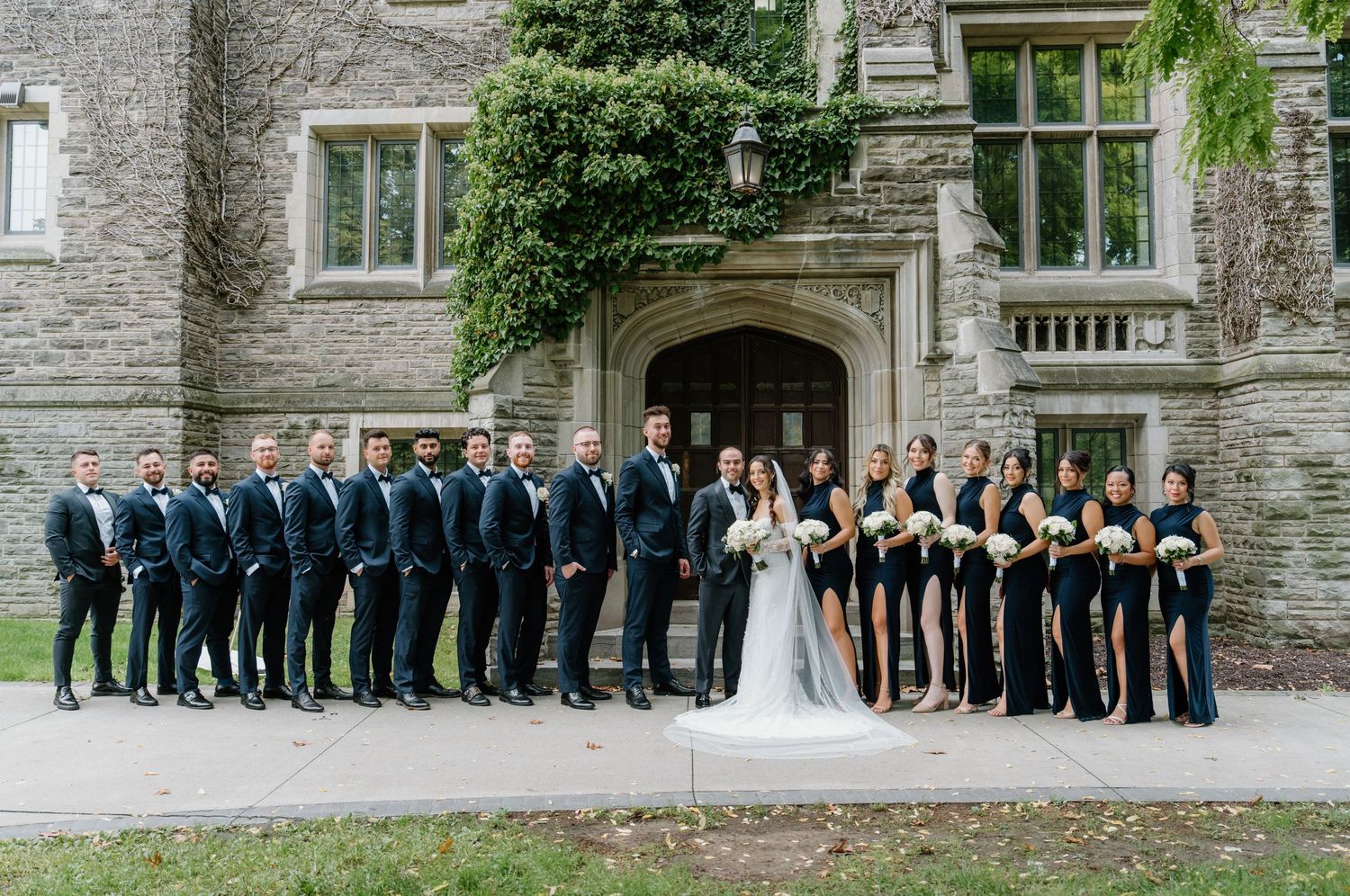 Wedding party in formal attire poses in front of stone building with elegant architecture.