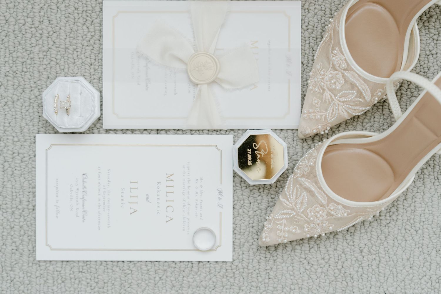 Flat lay of elegant wedding details in neutral tones with invitation, lace, rings, and flowers on textured background.