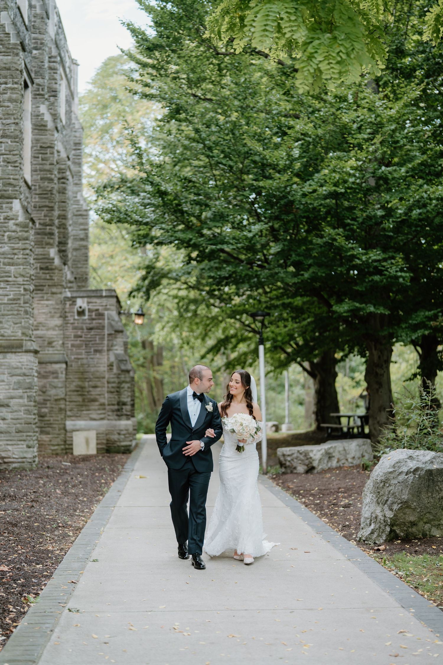 A couple in formal wedding attire walks along a tree-lined stone path at a historic venue.