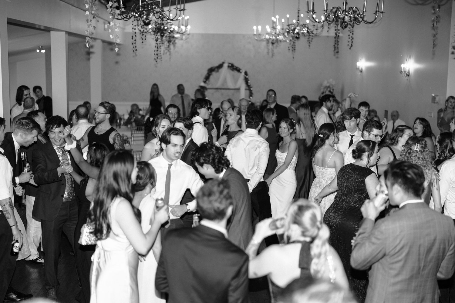Wedding guests dancing and celebrating on a crowded dance floor under crystal chandeliers at an elegant reception.