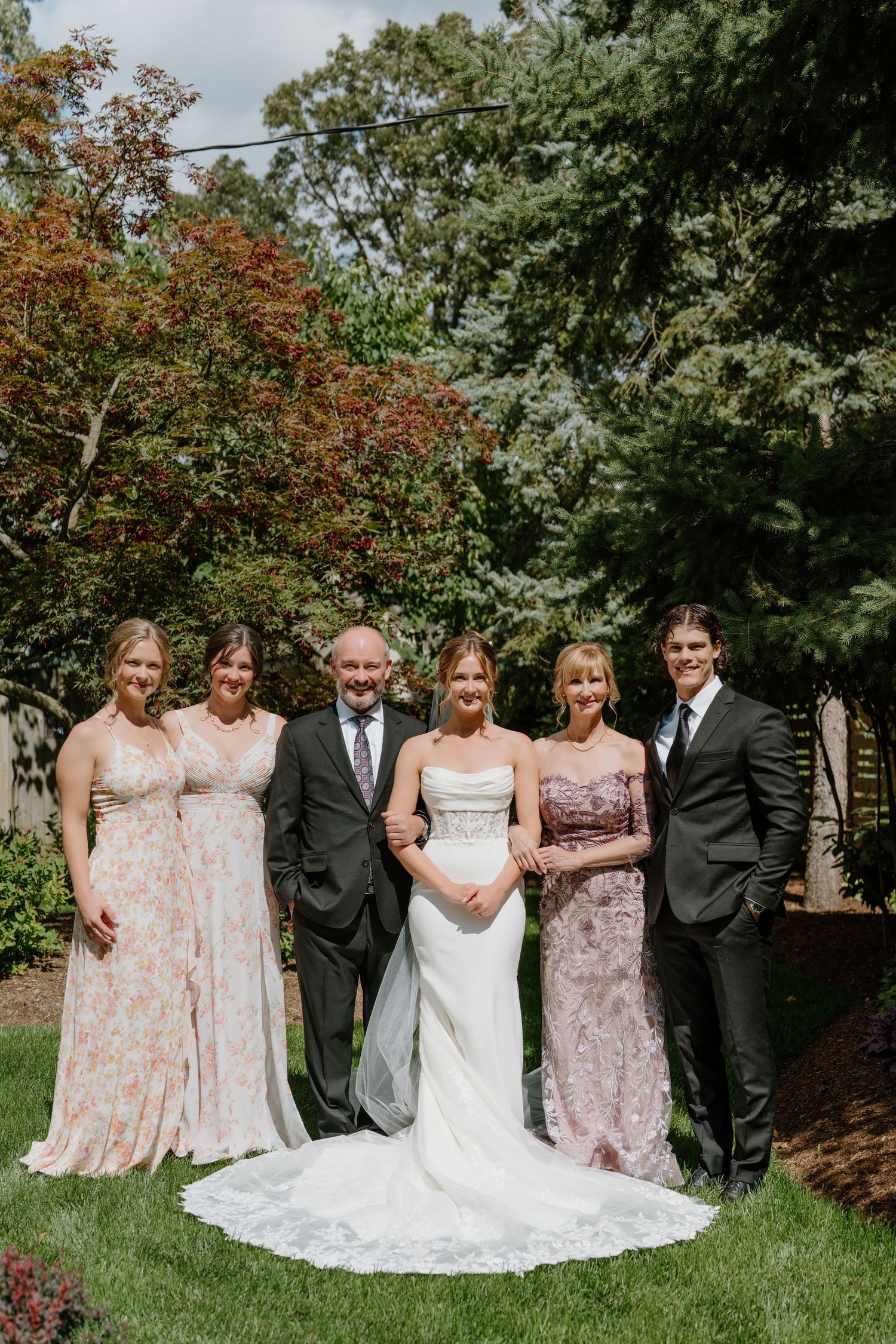 Wedding party poses outdoors in floral bridesmaid dresses and formal attire against lush green garden backdrop.