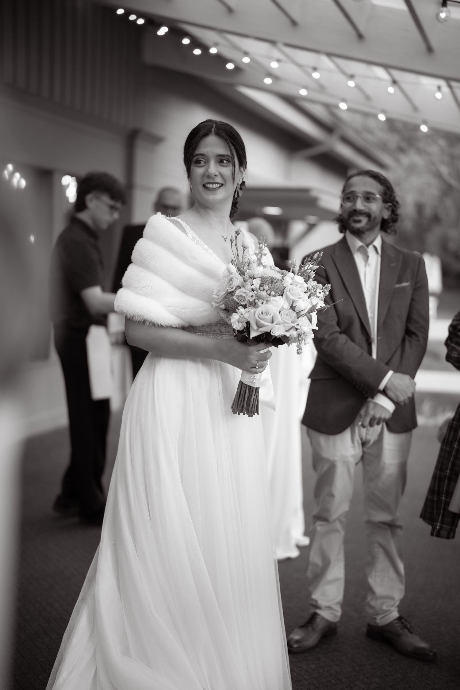 Wedding scene with bride in white dress and fur shawl holding a floral bouquet indoors.