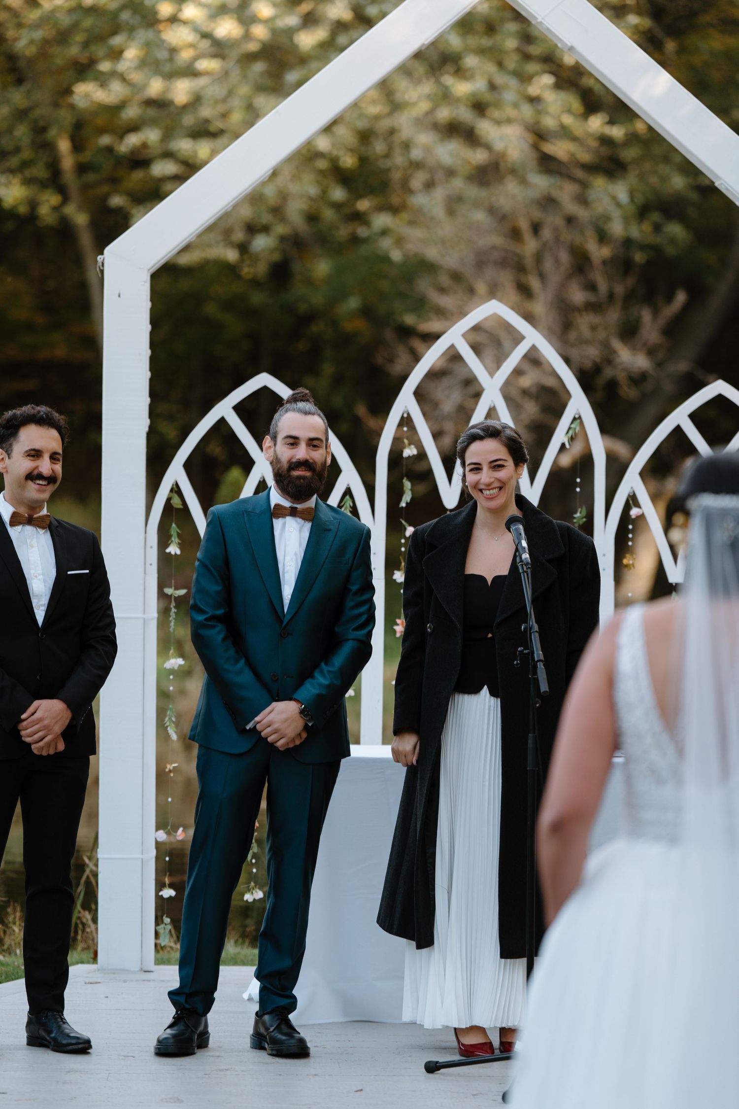 Wedding party standing under white chapel arch during outdoor ceremony.