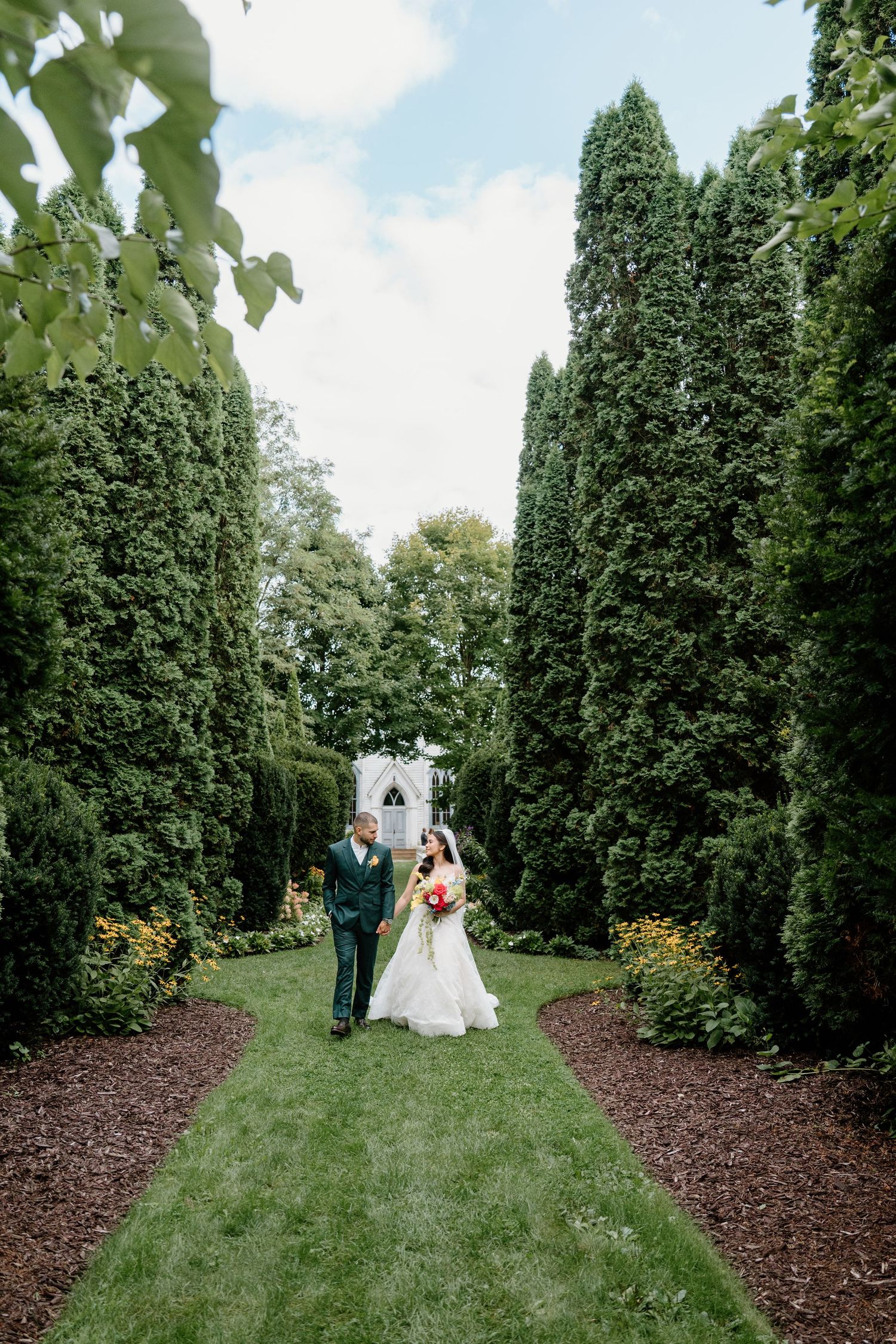 A wedding ceremony setup in a garden path lined with tall evergreen trees and mulched flower beds.