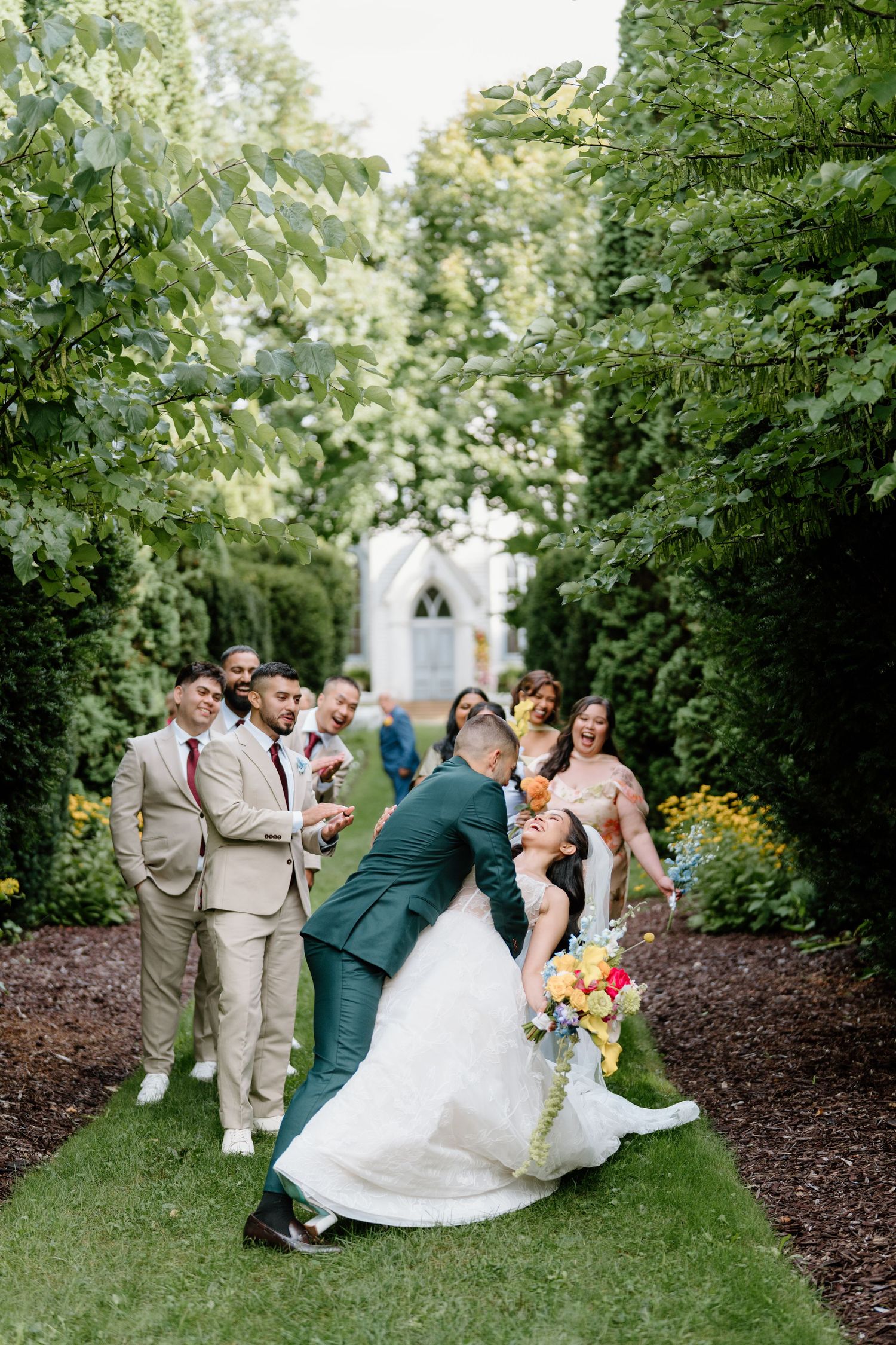 Wedding party poses together on a garden path with lush greenery and a chapel visible in the background.