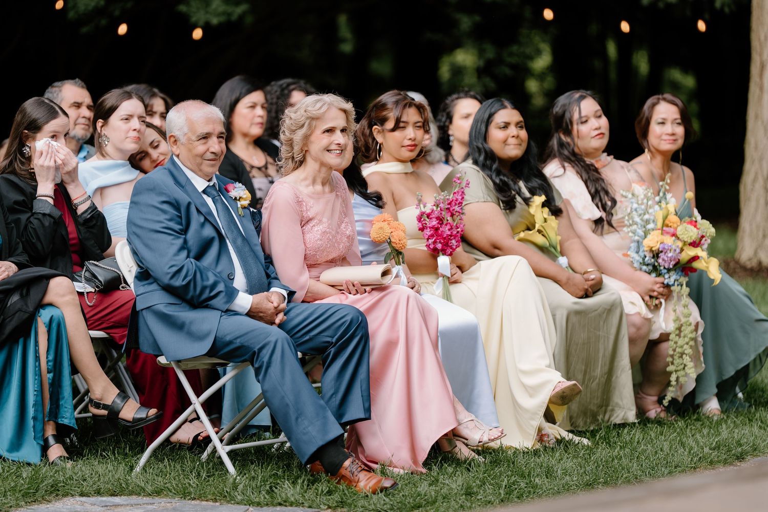 Rows of wedding attendees sitting on white chairs during an outdoor summer celebration.