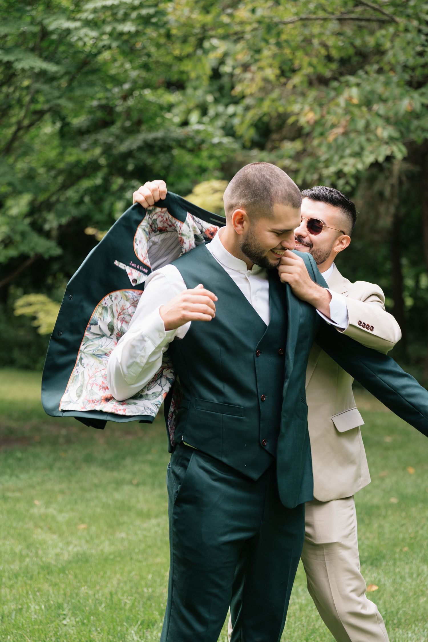 Two people in suits sharing a playful moment outdoors with green foliage in the background.