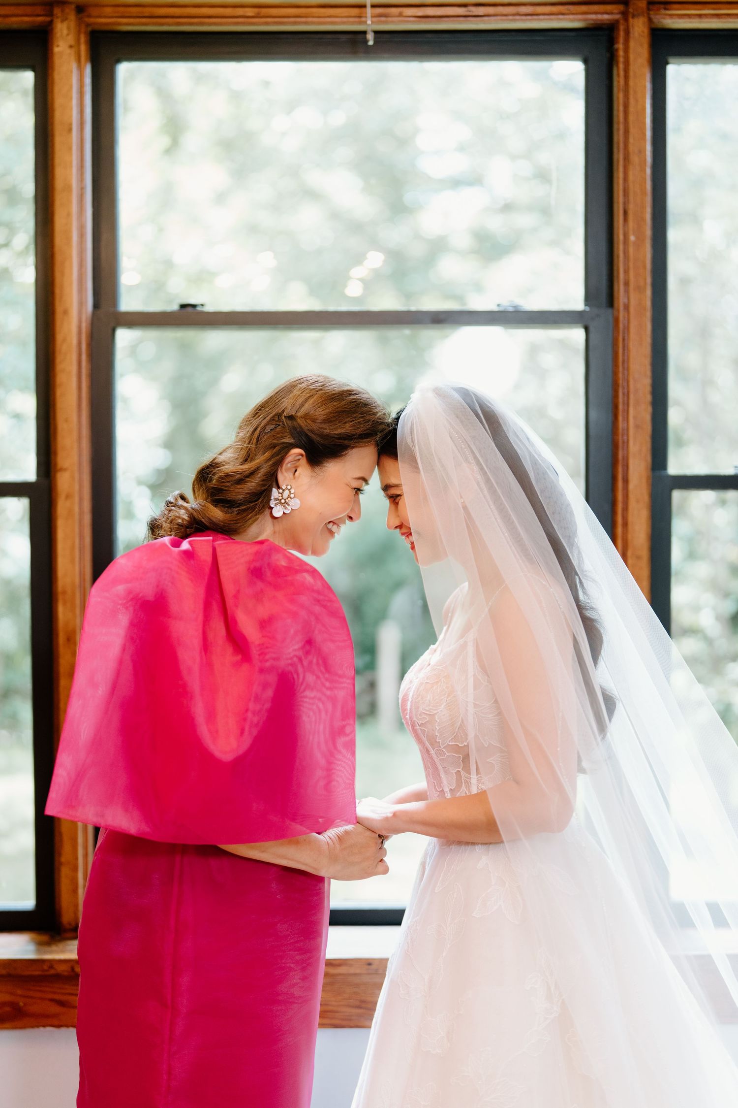 A person in bright pink embraces someone wearing a white wedding dress and veil in a sunlit room.