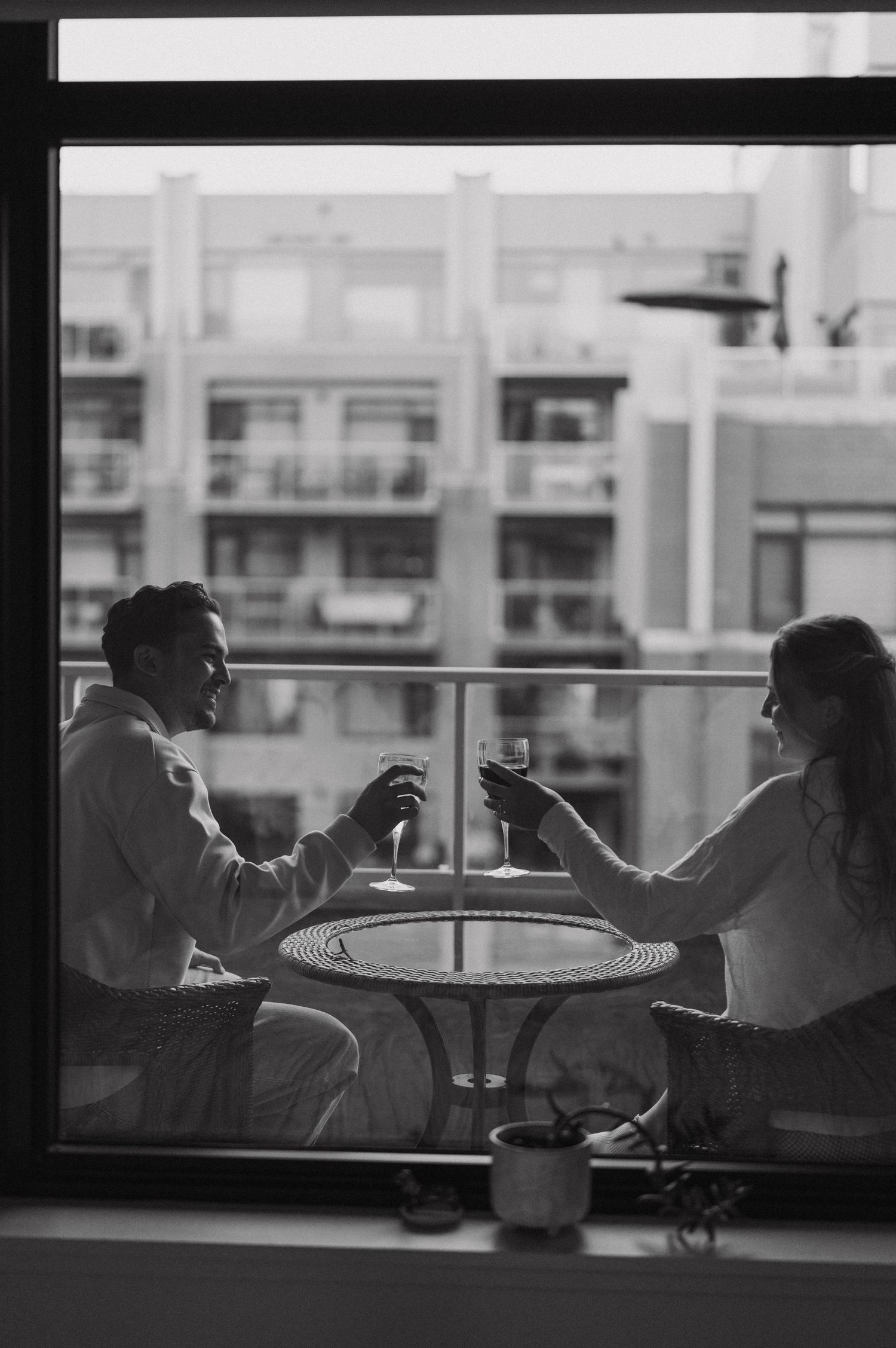 Two people share drinks on a balcony table silhouetted against an urban apartment building backdrop in black and white.
