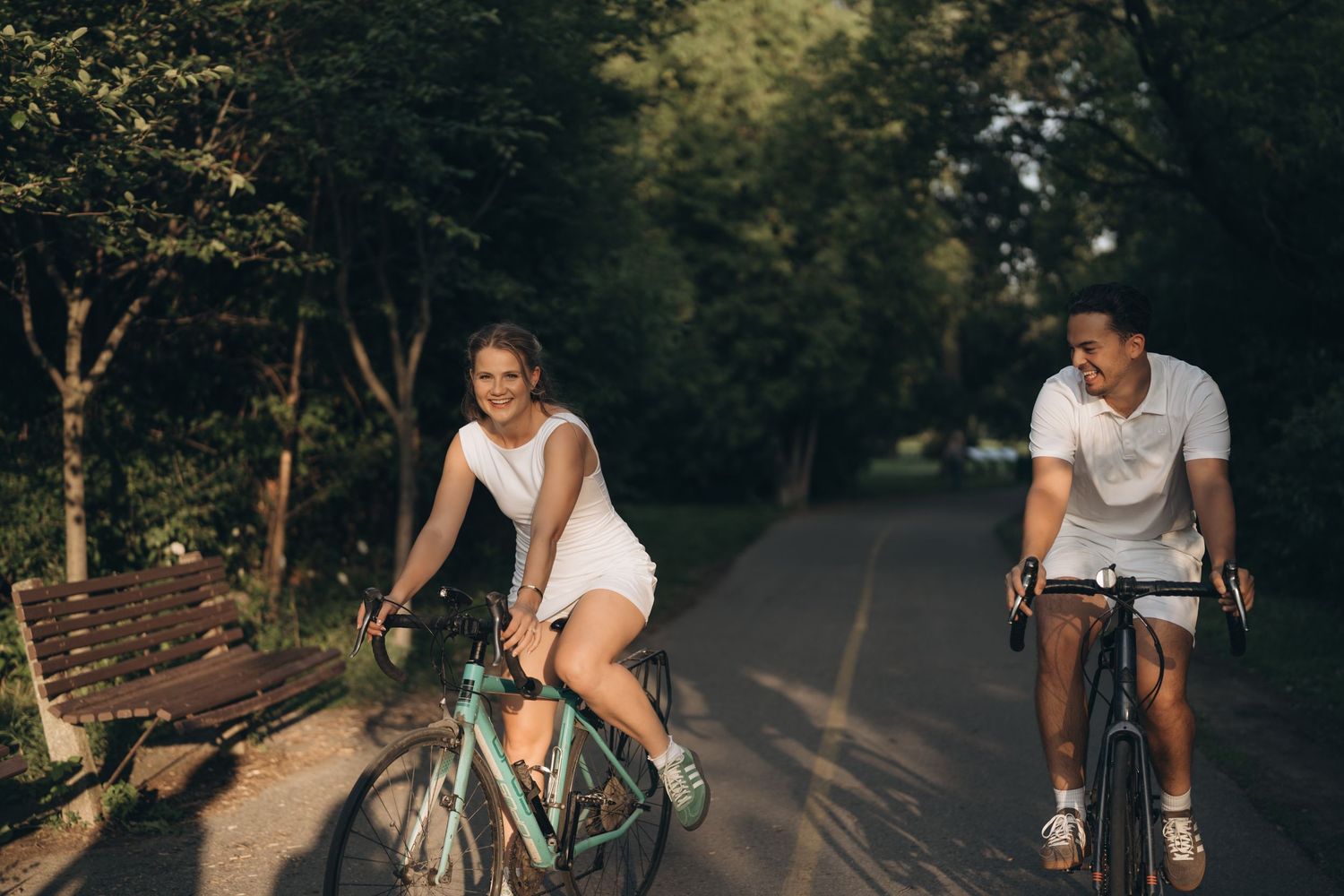 A couple enjoys a summer bike ride together on a tree-lined path in the late afternoon sunlight.