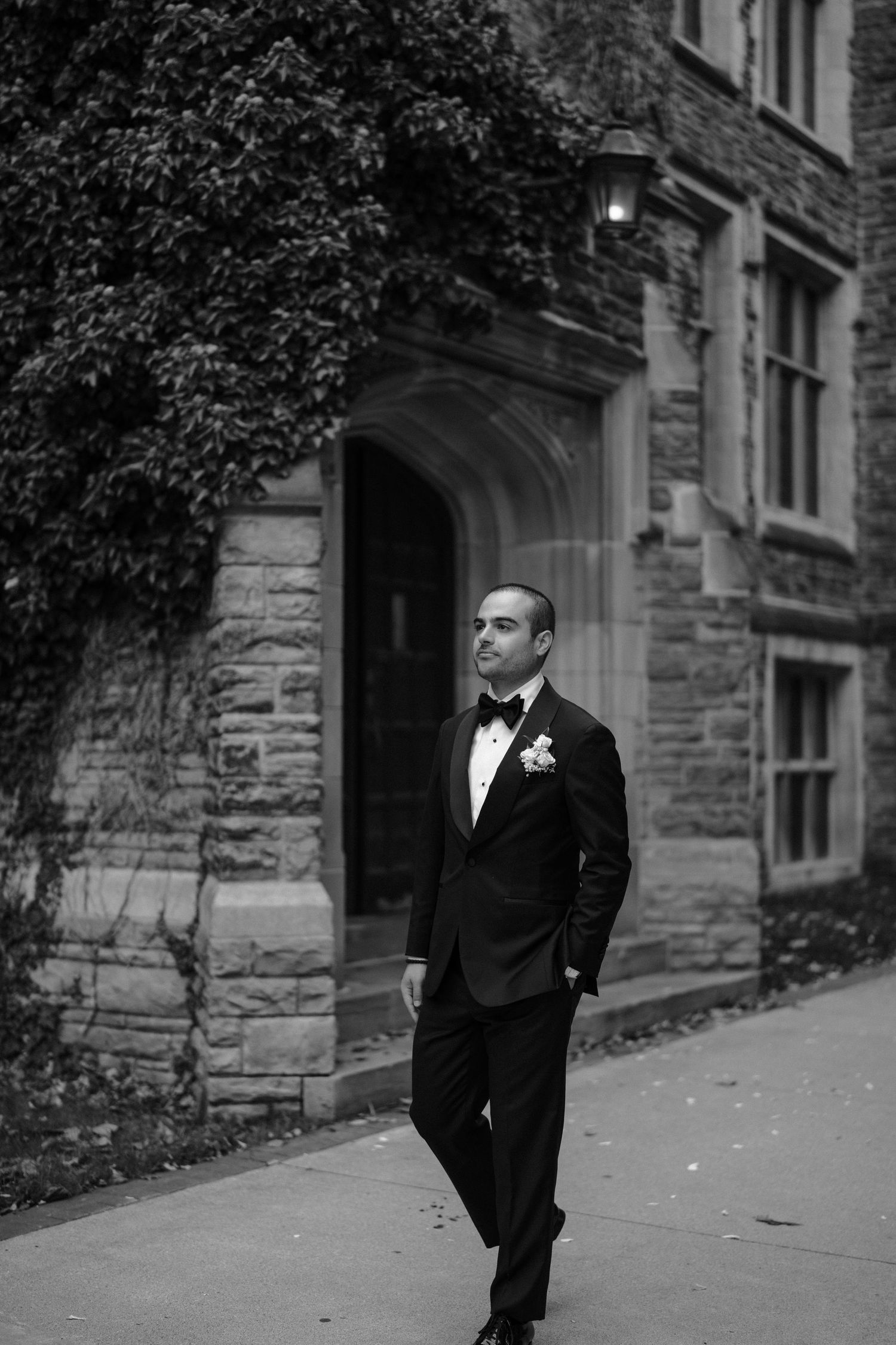 A groom in a classic black tuxedo poses thoughtfully against a stone building exterior.