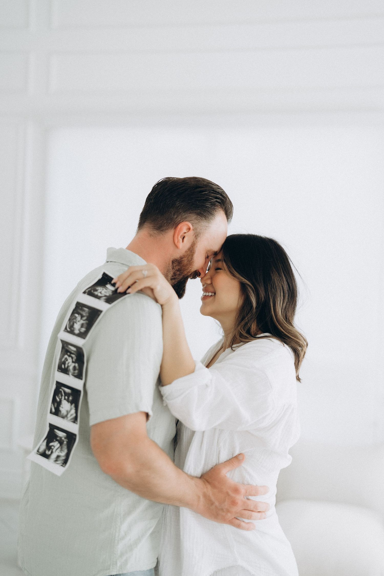 A couple in white clothing embraces intimately against a bright white wall with ultrasound photos displayed.