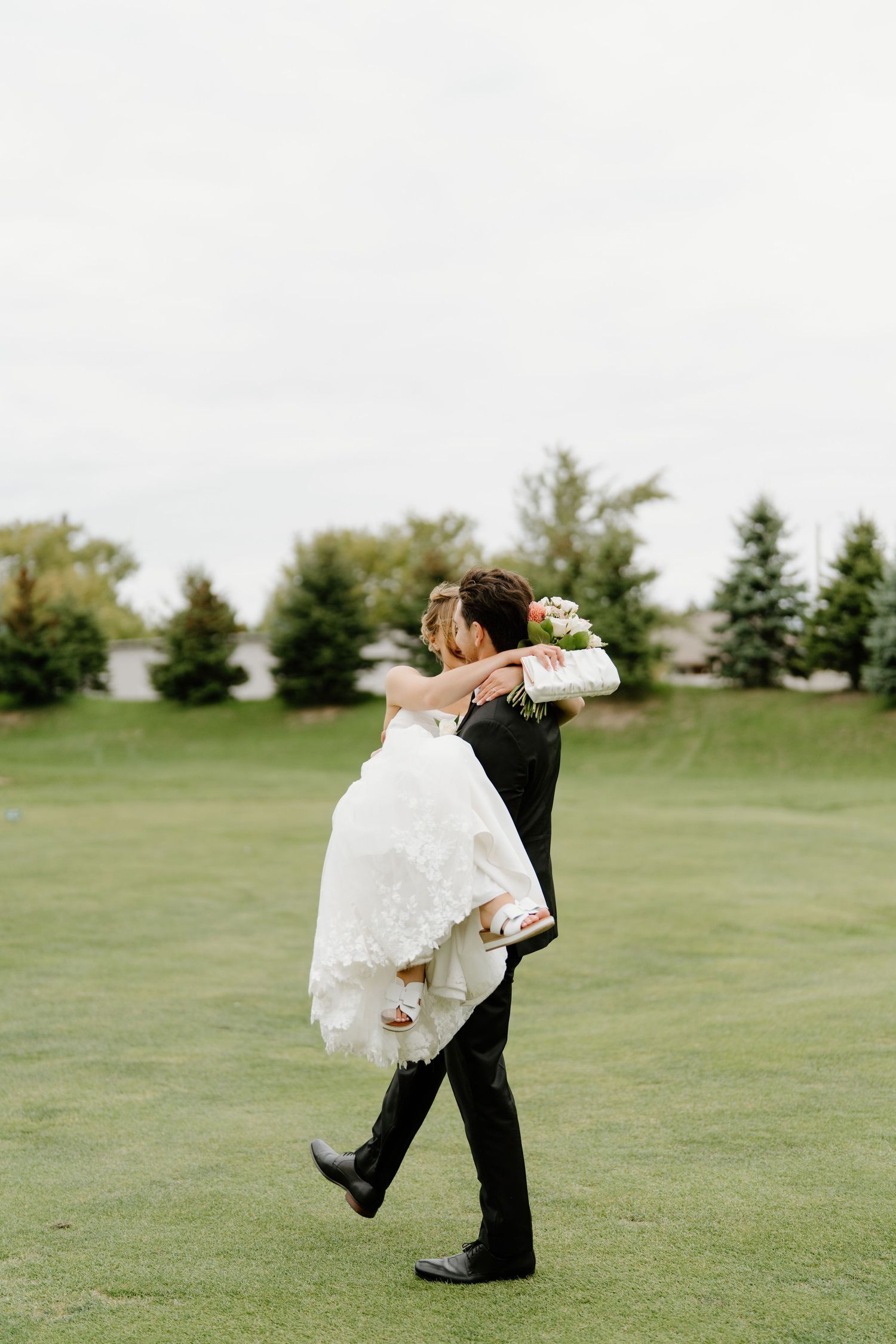 A couple in formal wedding attire dance and twirl together on a lush green lawn with trees in the background.