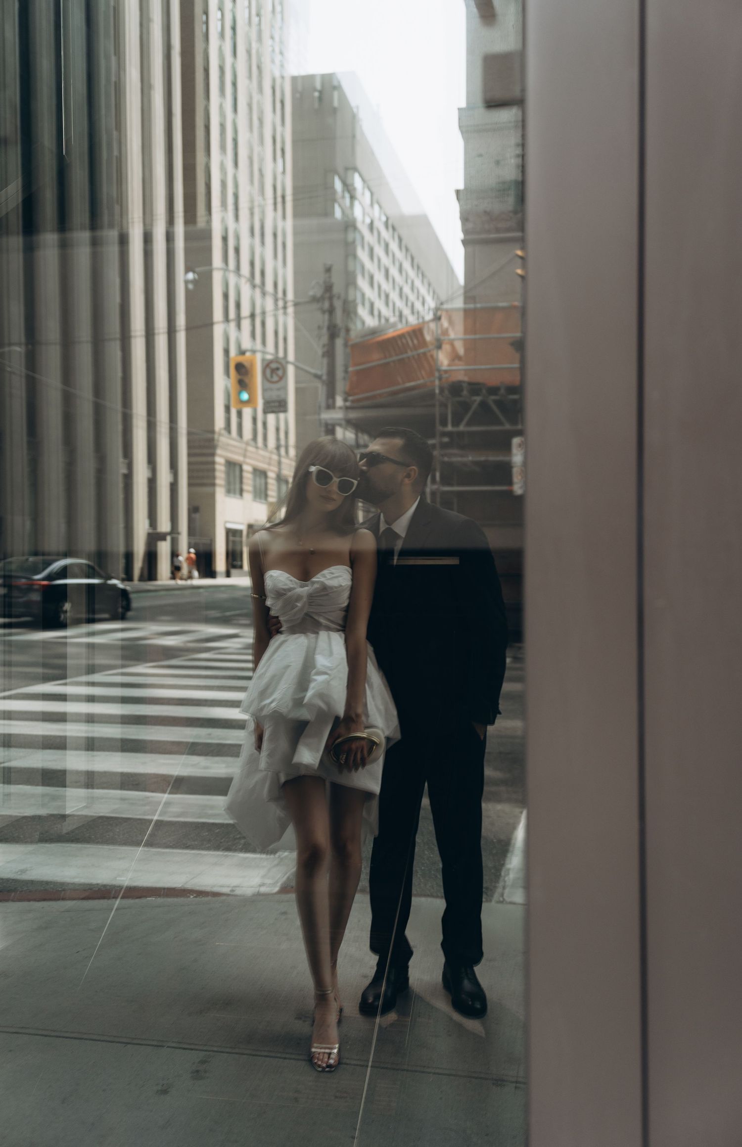 A couple in formal wear walks together along a downtown sidewalk reflected in mirrored windows.