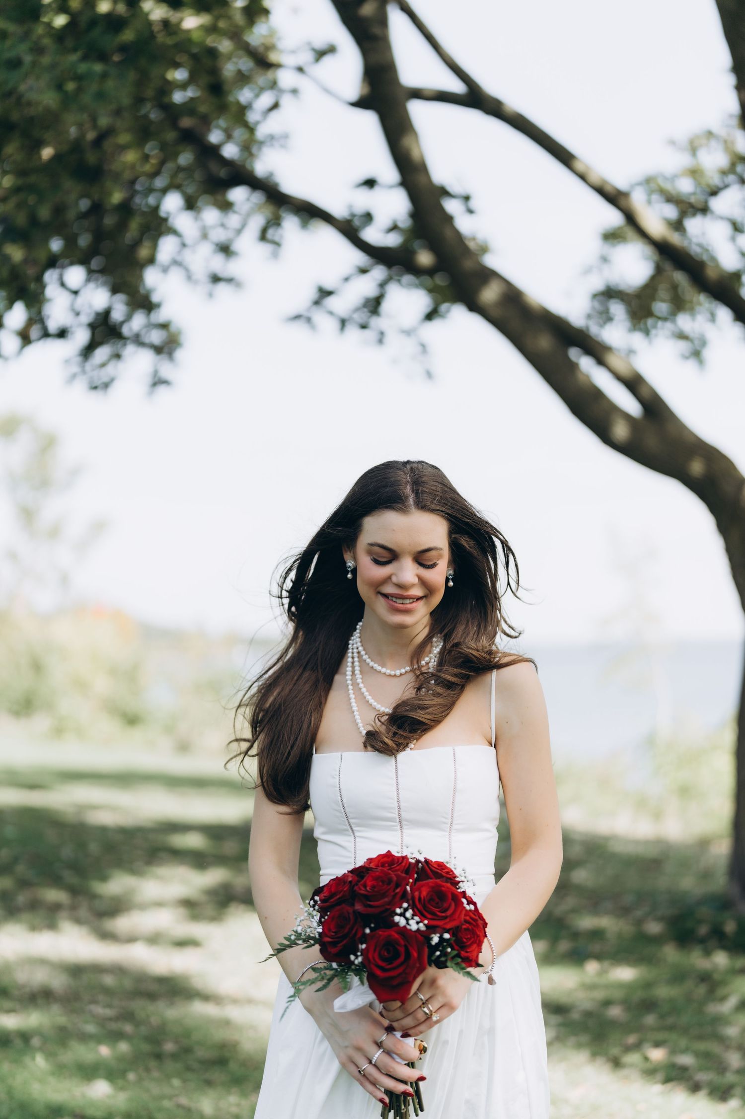 Bride in white wedding dress holding deep red bouquet of roses outdoors under trees.