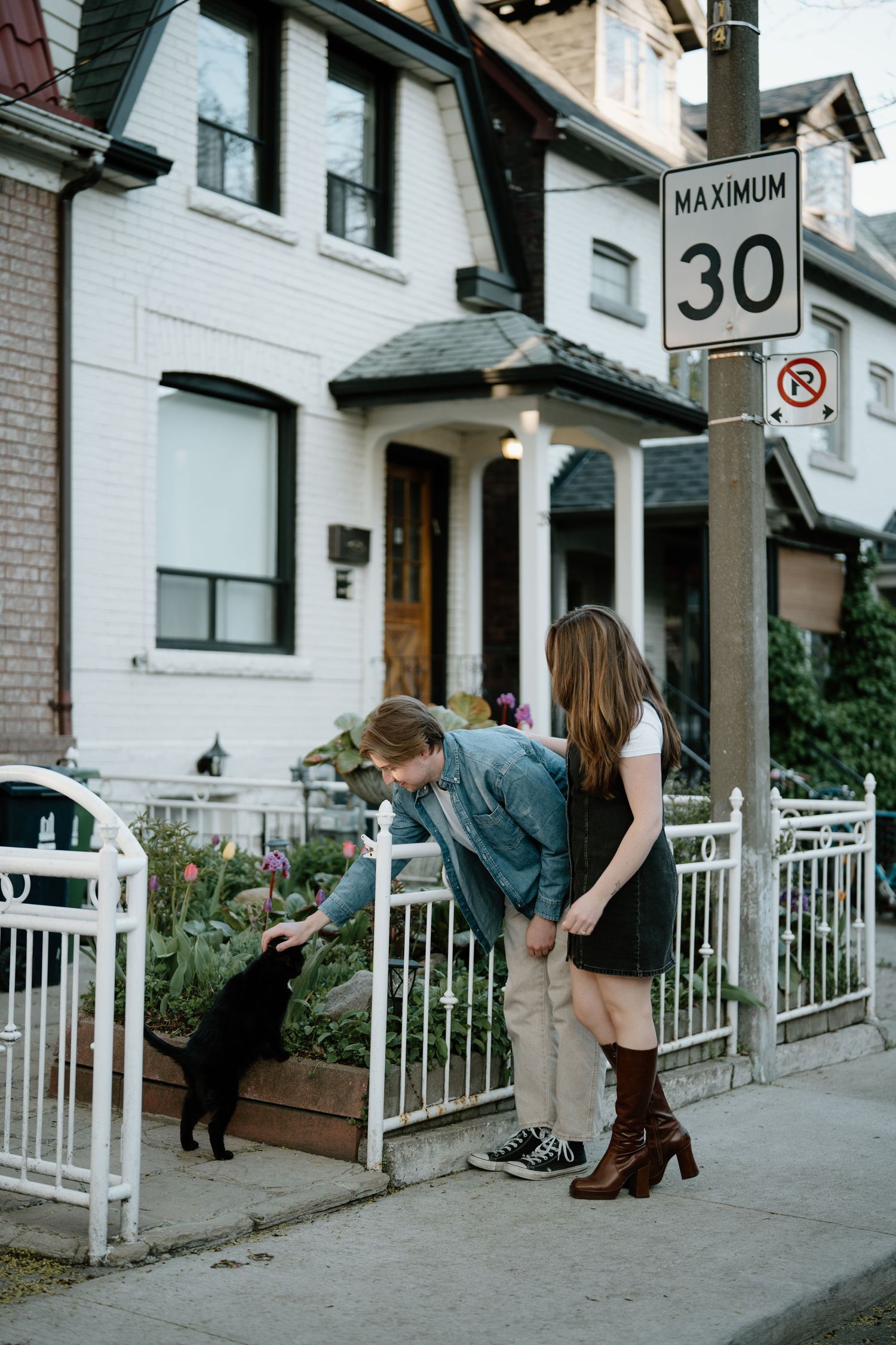Two people petting a black cat on a sidewalk beside a white house with a 30km speed limit sign.