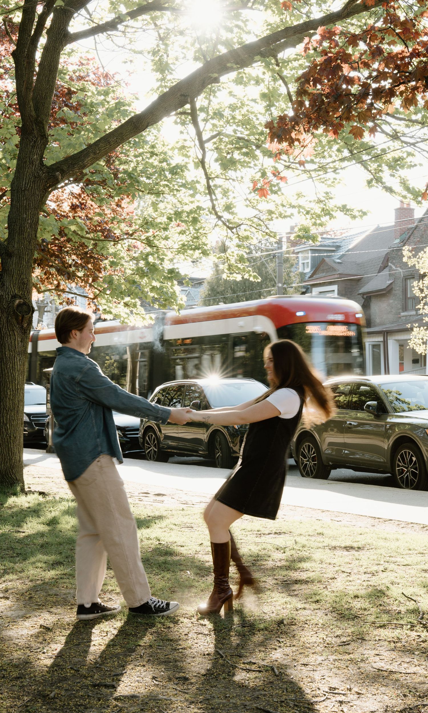 A couple dances playfully together on a tree-lined street during golden hour as a bus passes by in the background.