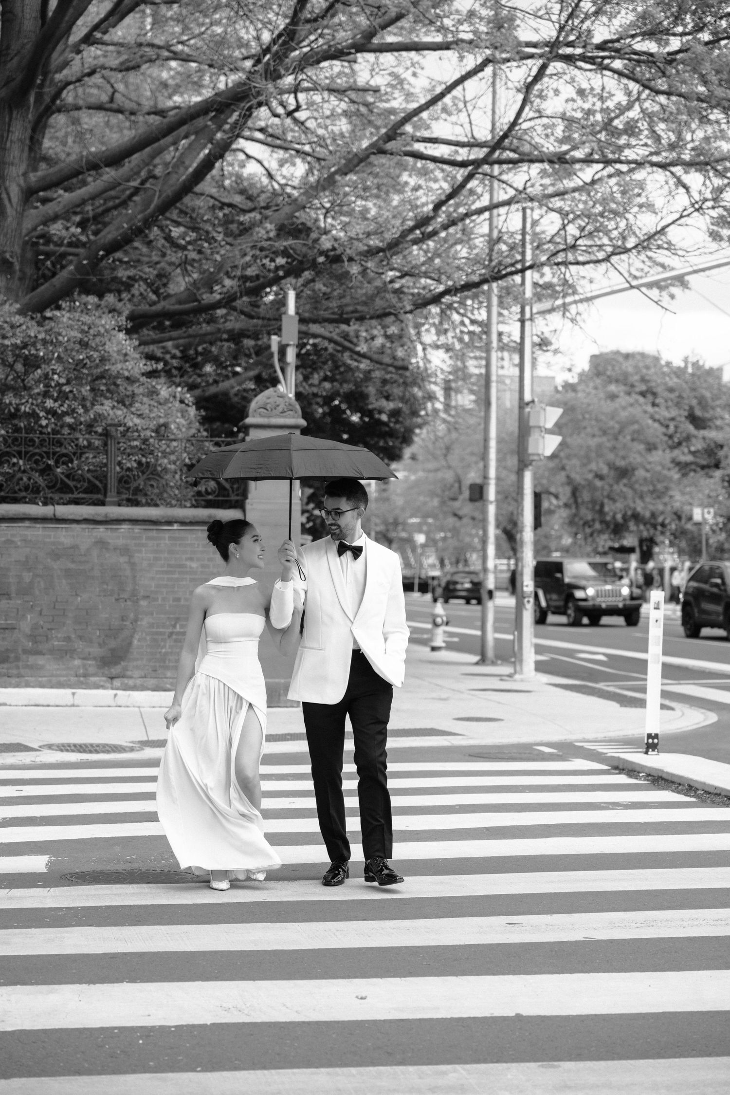 Black and white photo of a couple in formal attire crossing a city street together on a crosswalk under leafy trees.