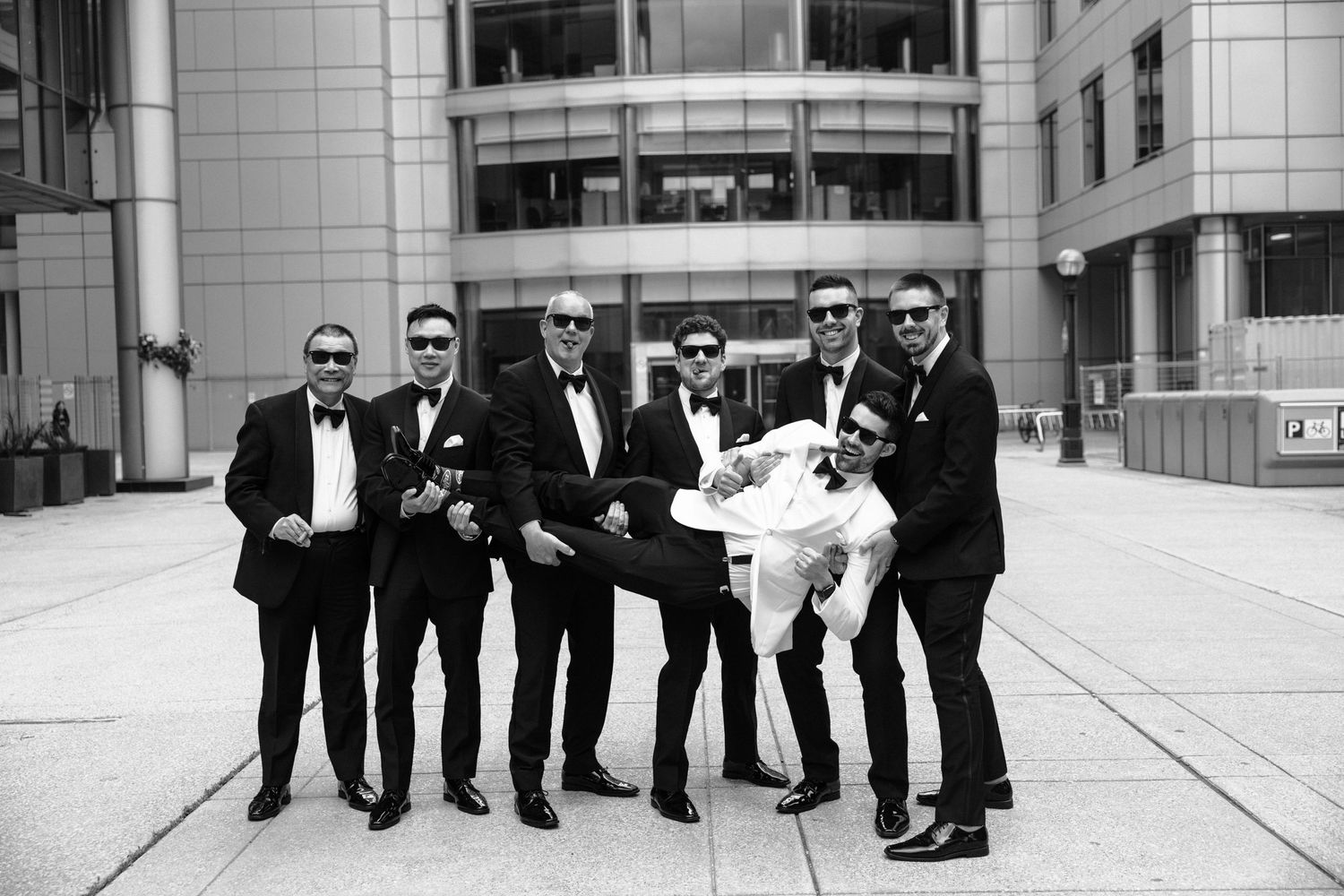 A black and white photo shows a group of men in suits and sunglasses posing together outside a modern building.