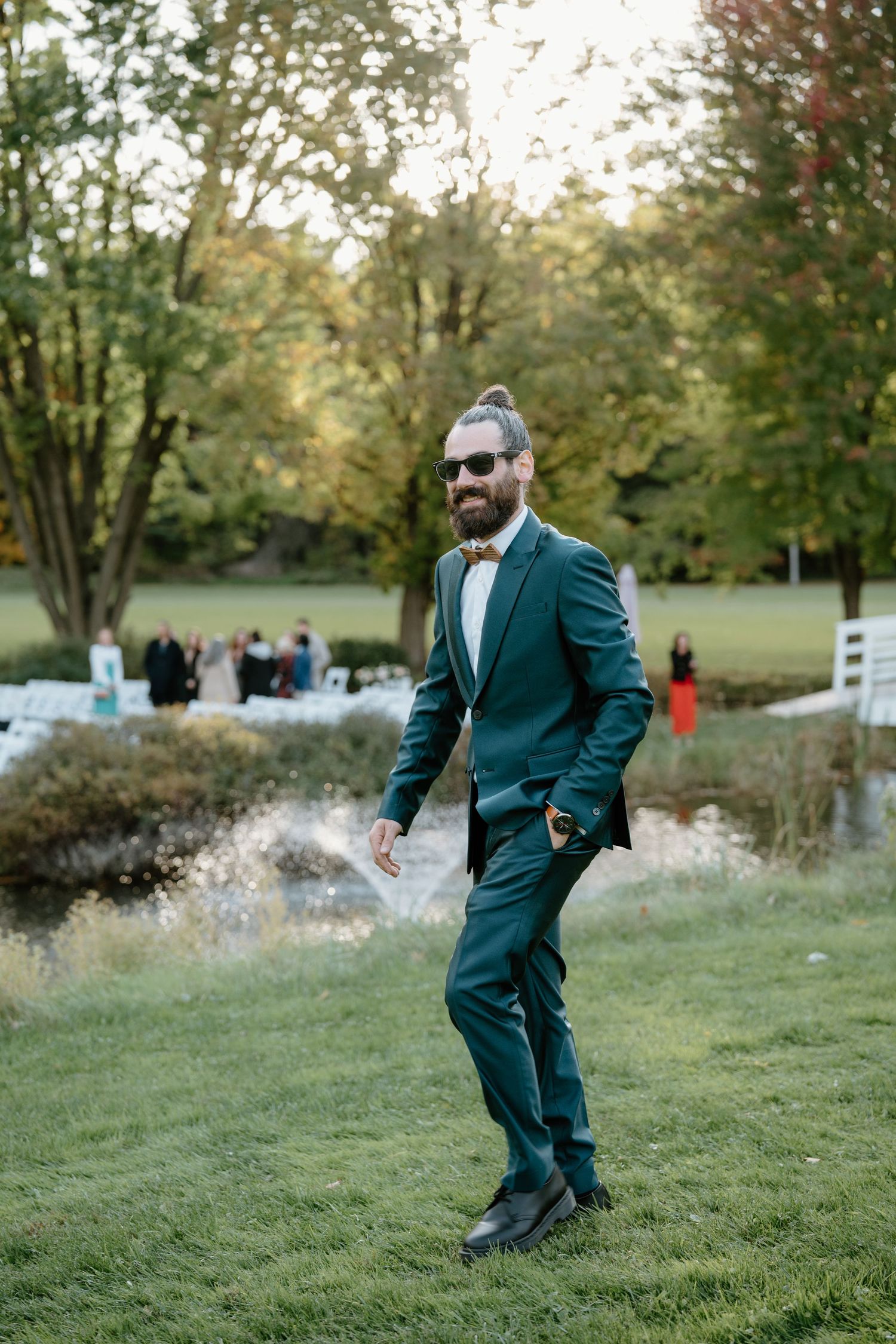 Groom in teal suit walking on grass with white fence and autumn trees in background.