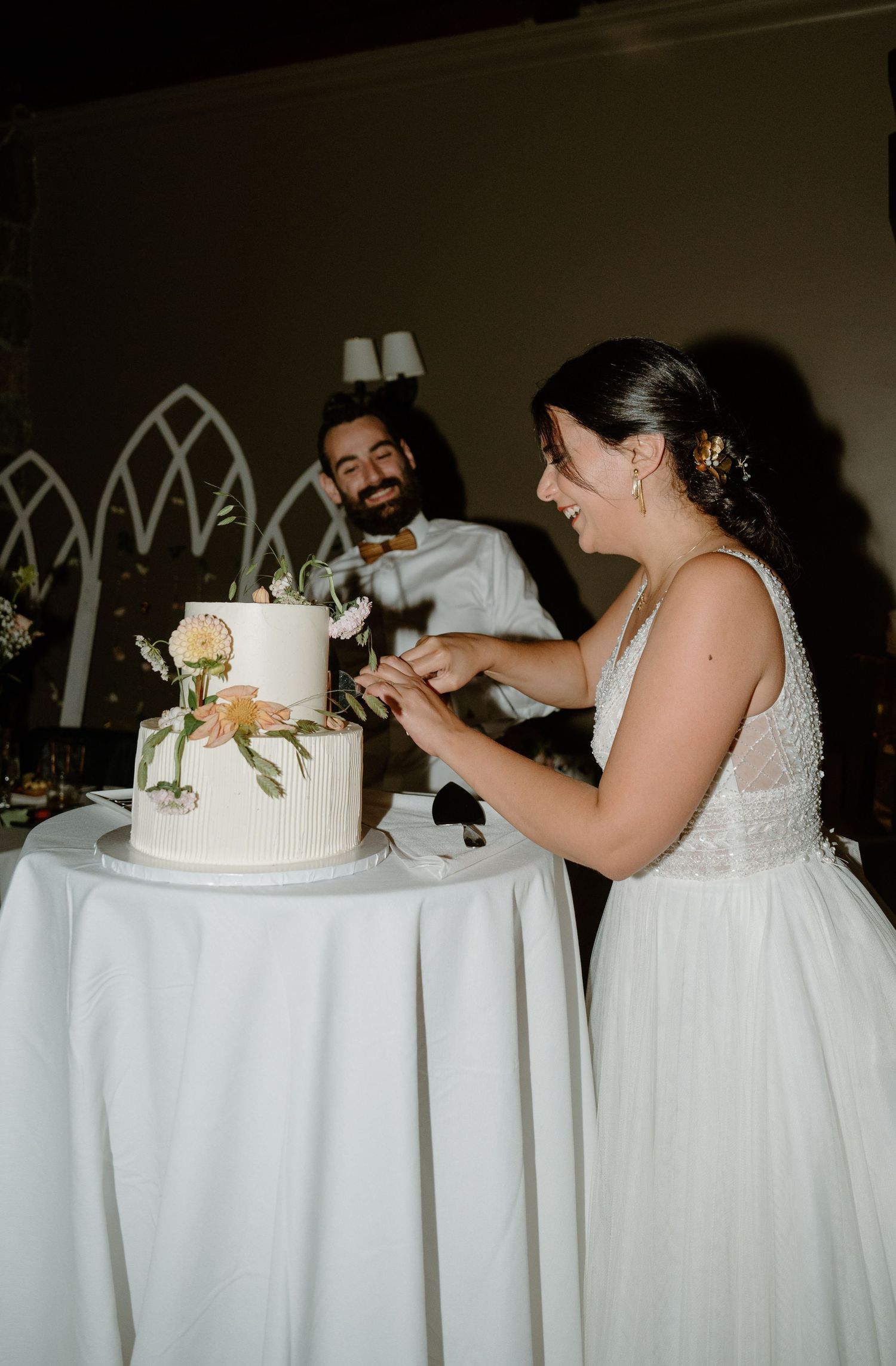 A couple laughs together while cutting their two-tiered white wedding cake at their reception.