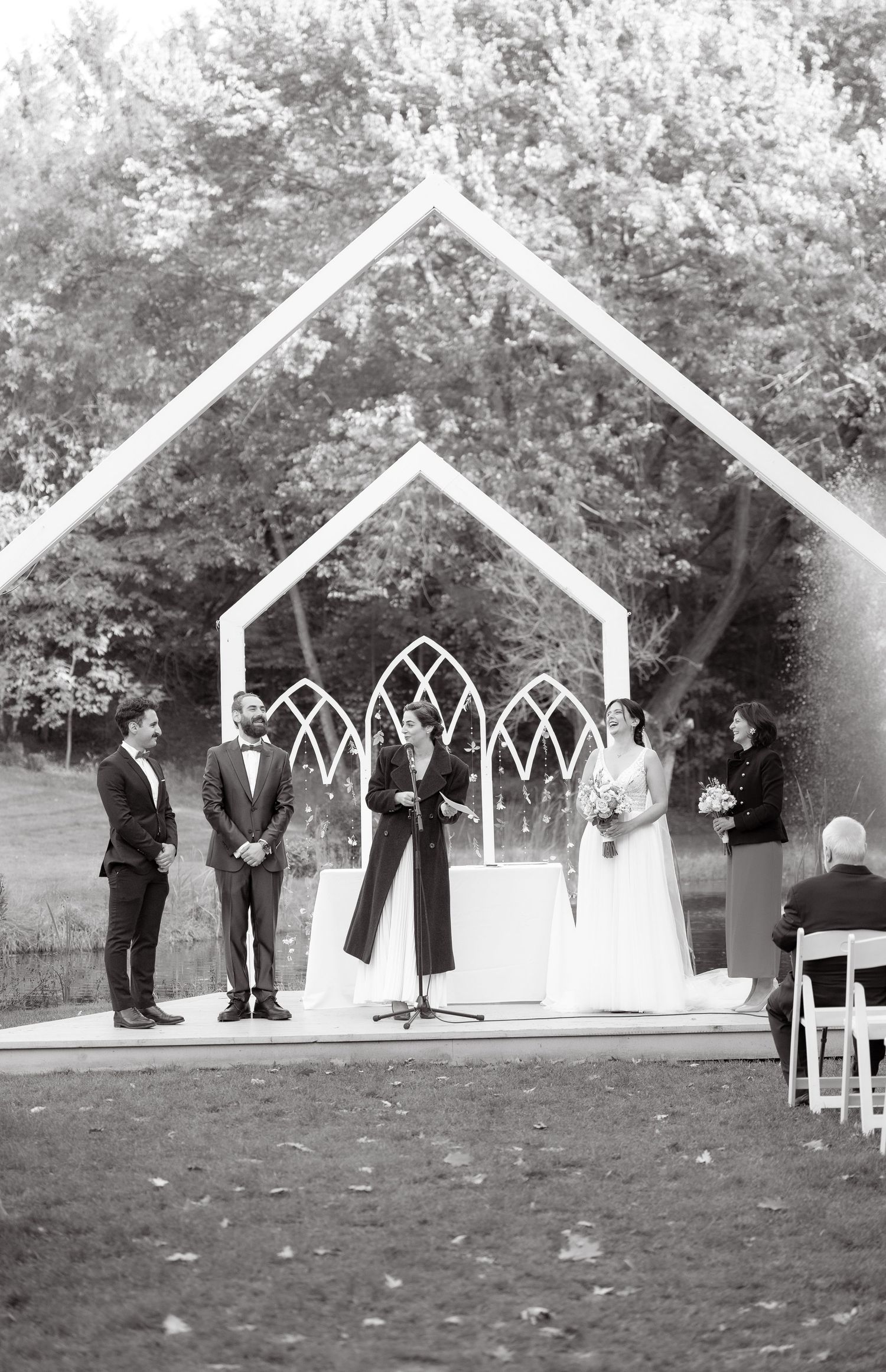 Black and white photo of outdoor wedding ceremony under geometric white archway.