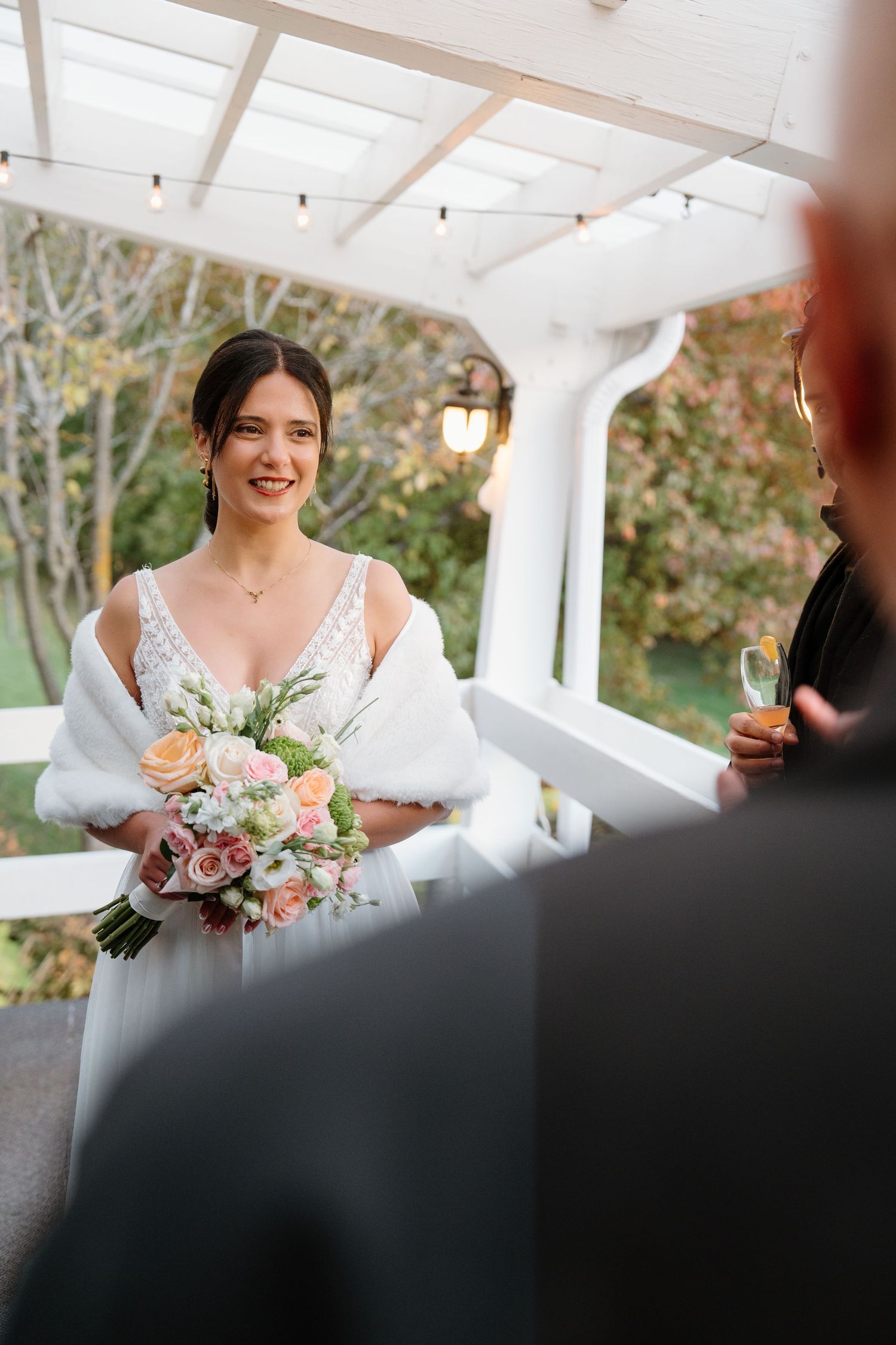 A bride holding a pink and peach bouquet stands under a white covered porch.