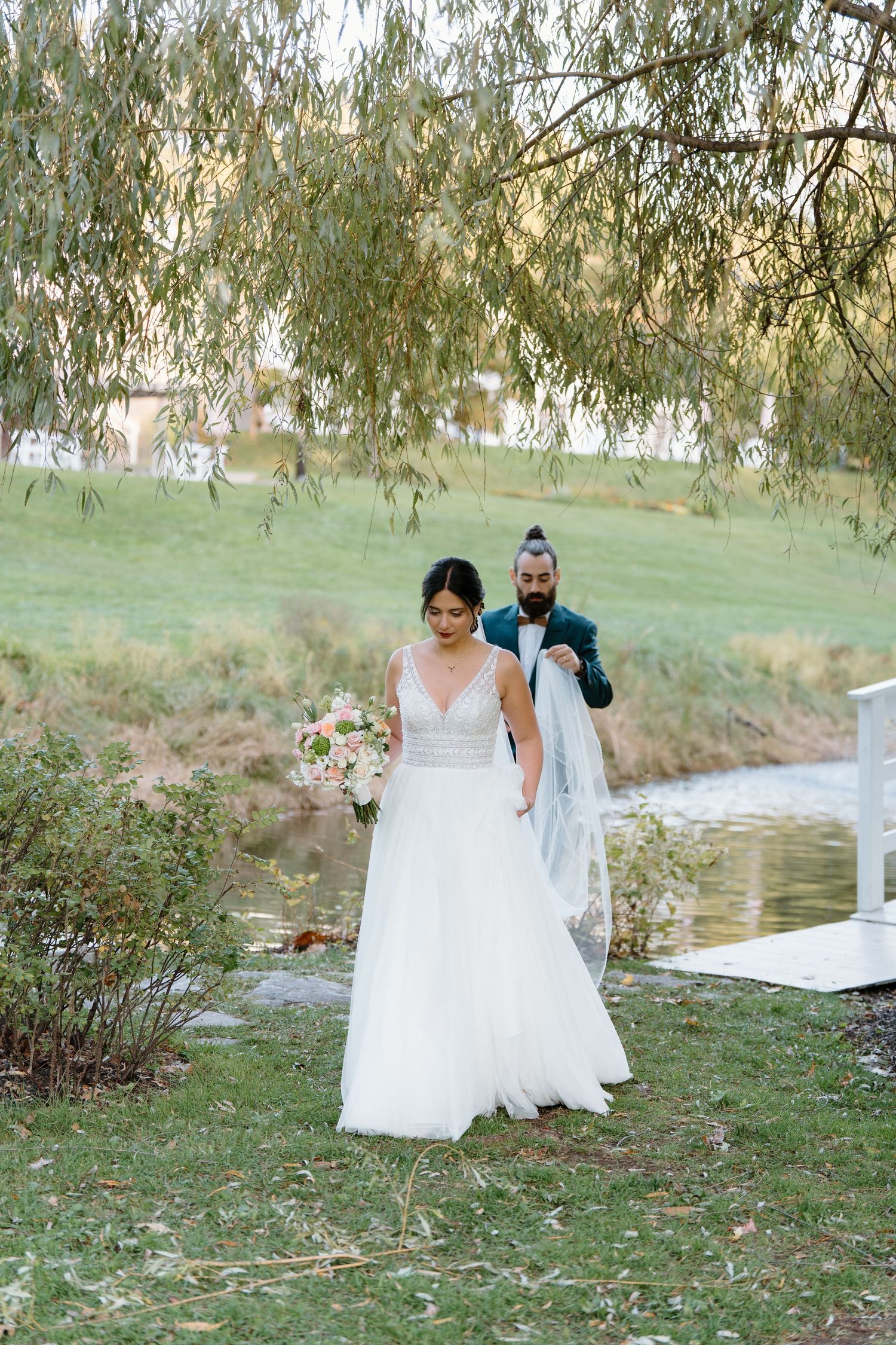 Bride in white gown walking beneath willow trees while holding bouquet during outdoor wedding.